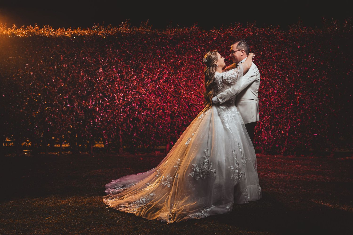 Novios bailan en escenario oscuro; fondo de luces rojas brillantes (posiblemente público). Boda.