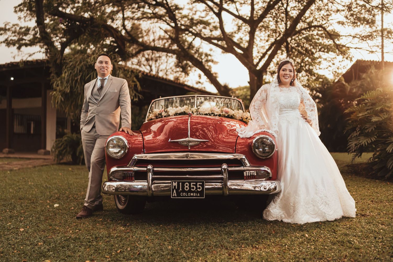 Novios posan elegantes junto a coche clásico Chevrolet rojo en jardín; ella vestido blanco, él traje gris. Boda.