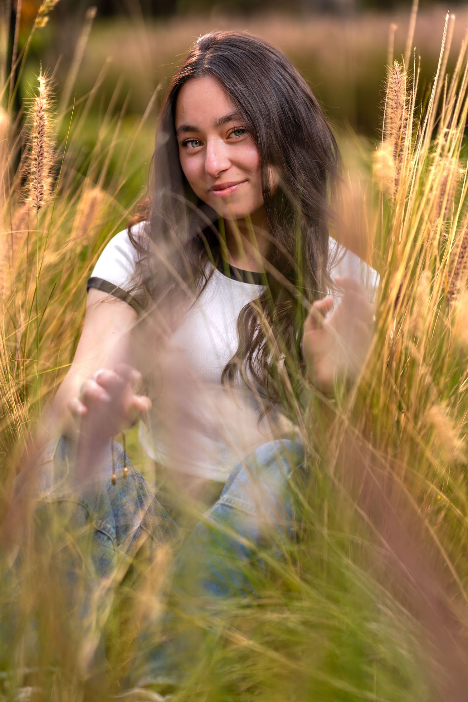 Joven mujer con top blanco entre pasto alto dorado extiende su mano a cámara; mirada suave. Sesión fotos.