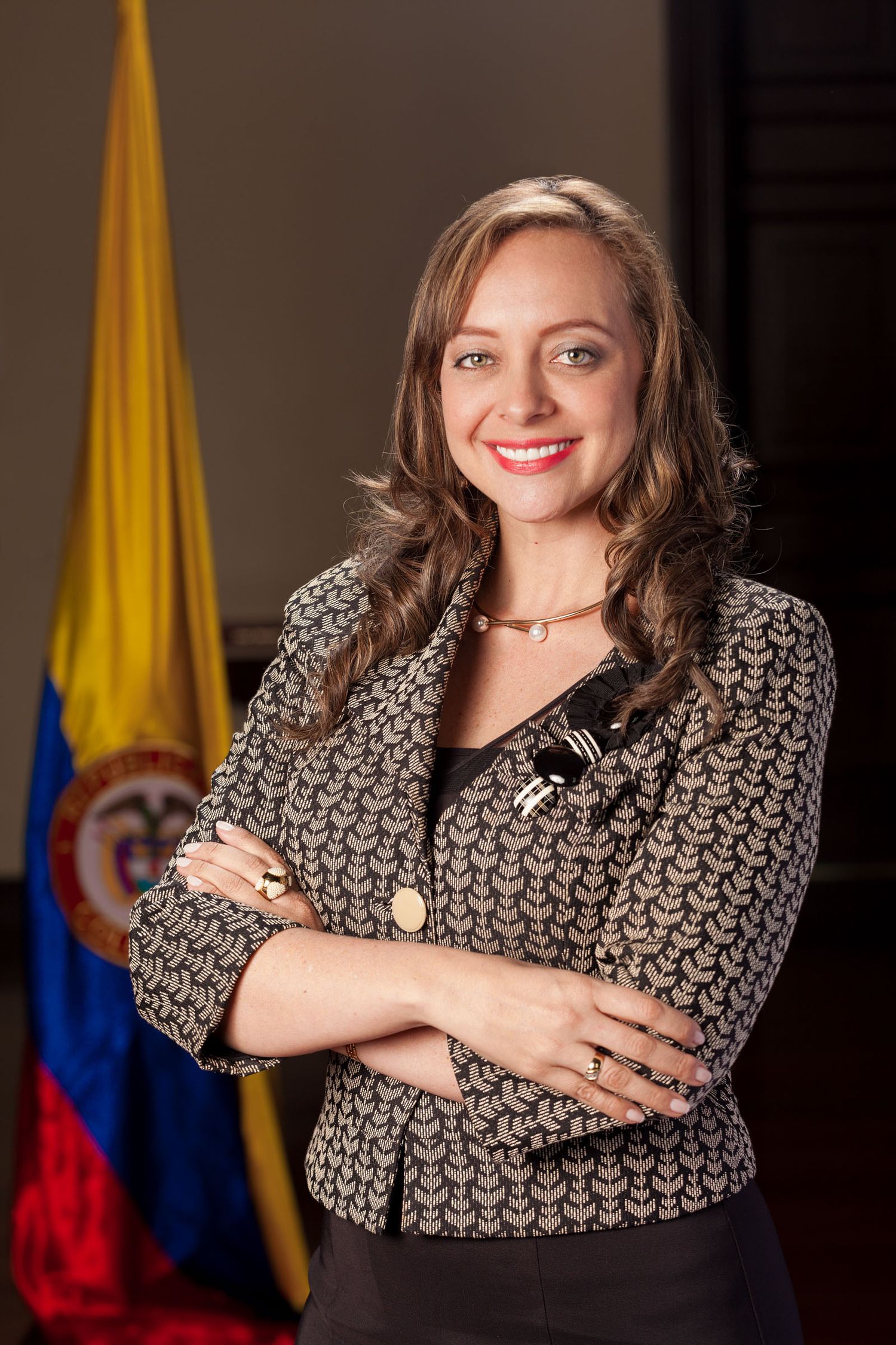 Mujer profesional sonriente con blazer estampado y brazos cruzados; bandera de Colombia al fondo. Retrato Ana Paola Agudelo.