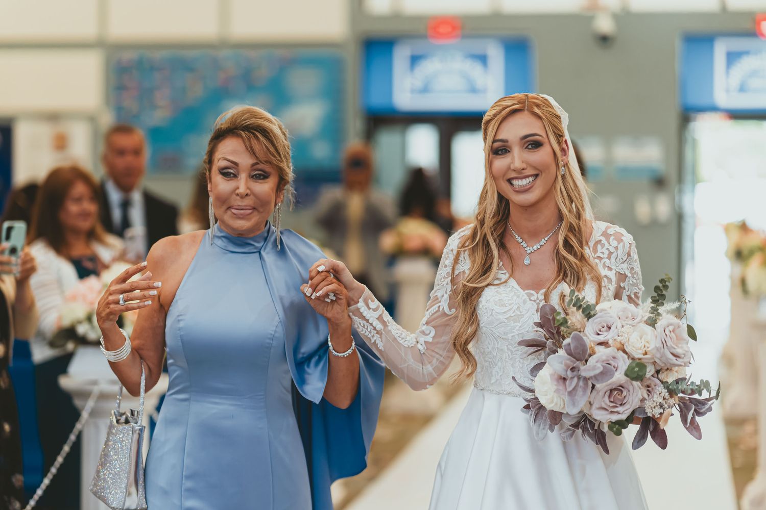 Novia sonriente con ramo camina del brazo de su madre (vestido azul) por pasillo iglesia. Entrada nupcial.