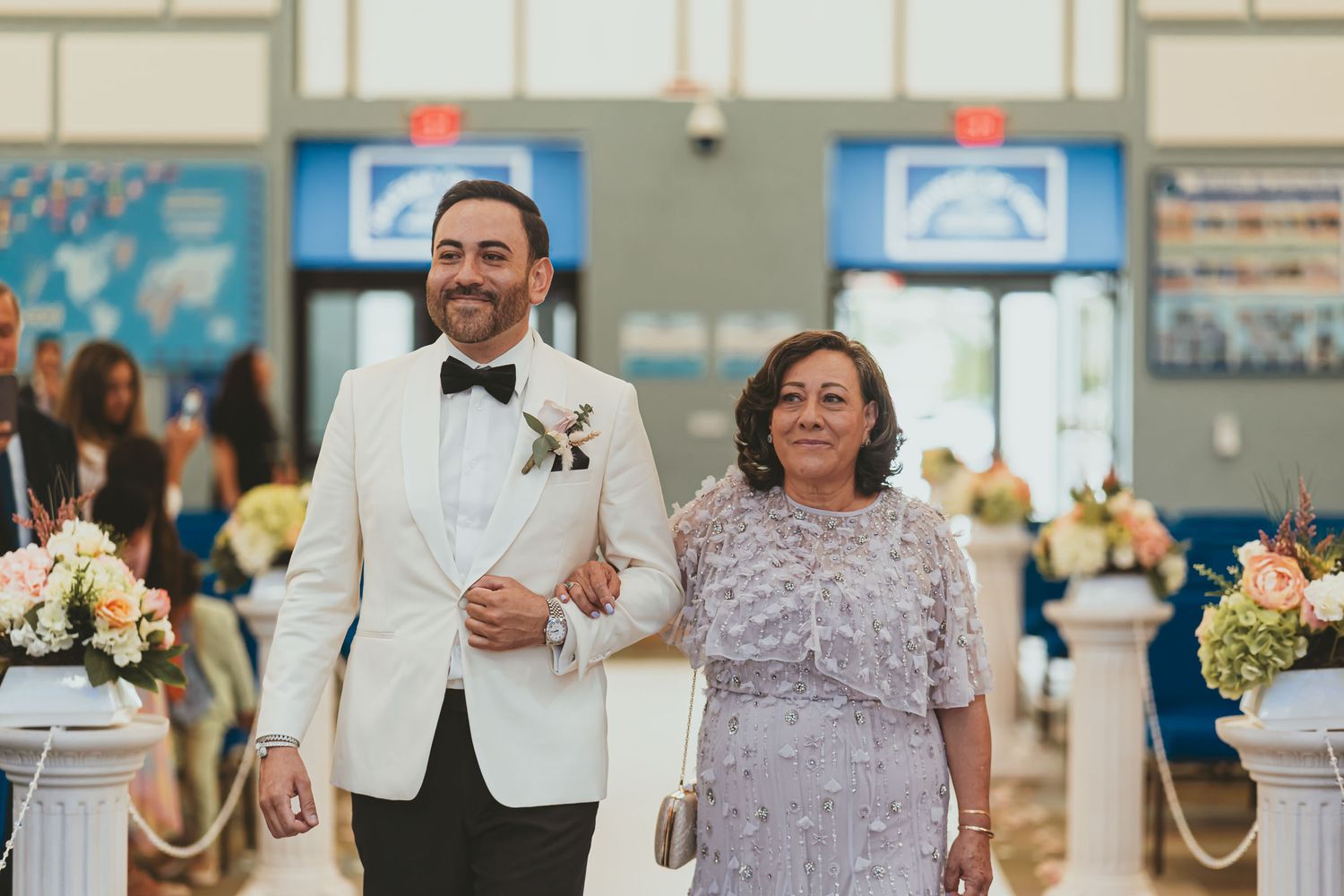 Novio sonriente (traje blanco) camina del brazo de su madre (vestido lavanda) por pasillo iglesia. Boda.