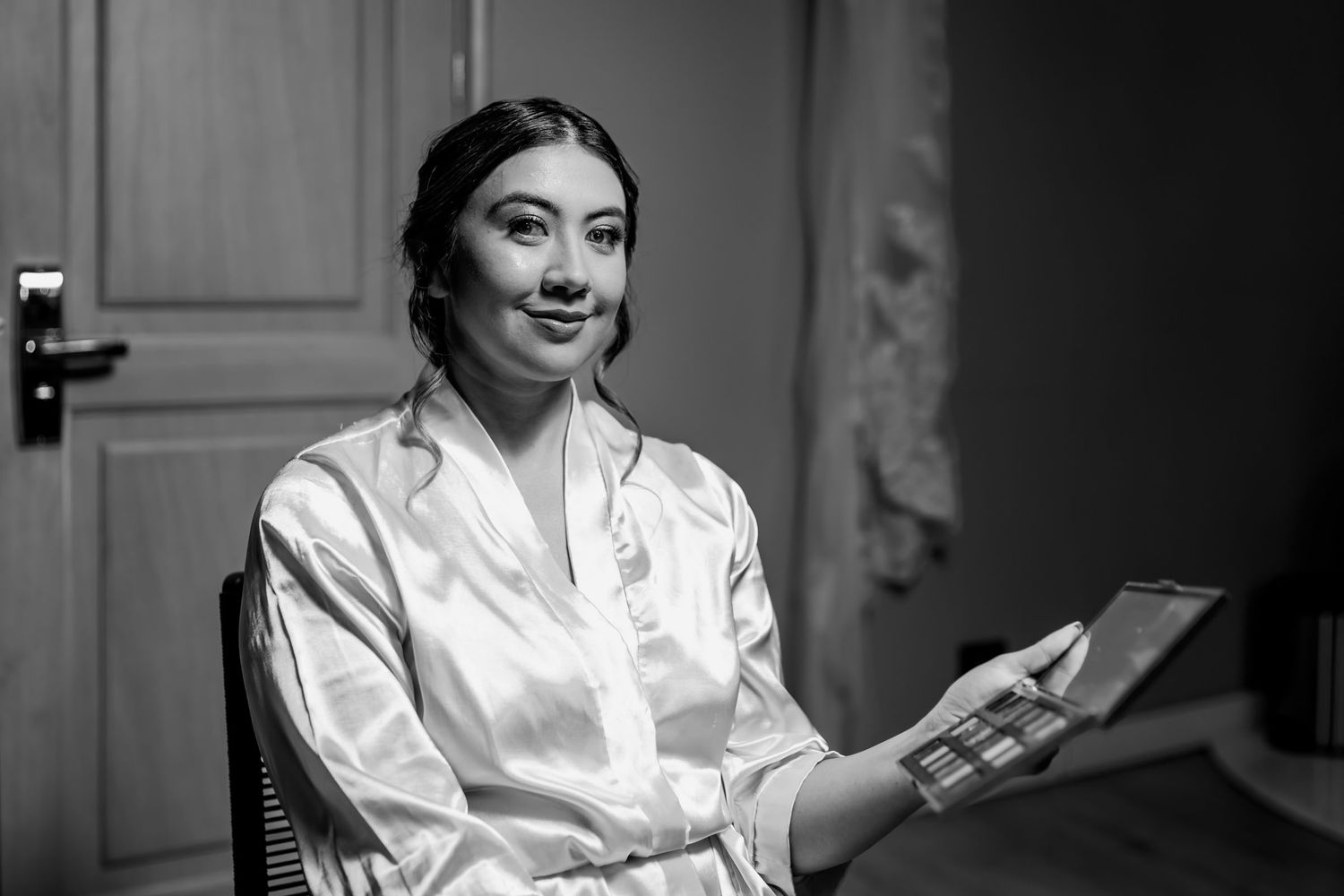 Foto B&W: Novia en bata de satín sostiene paleta de maquillaje durante preparación de boda; vestido colgado al fondo.