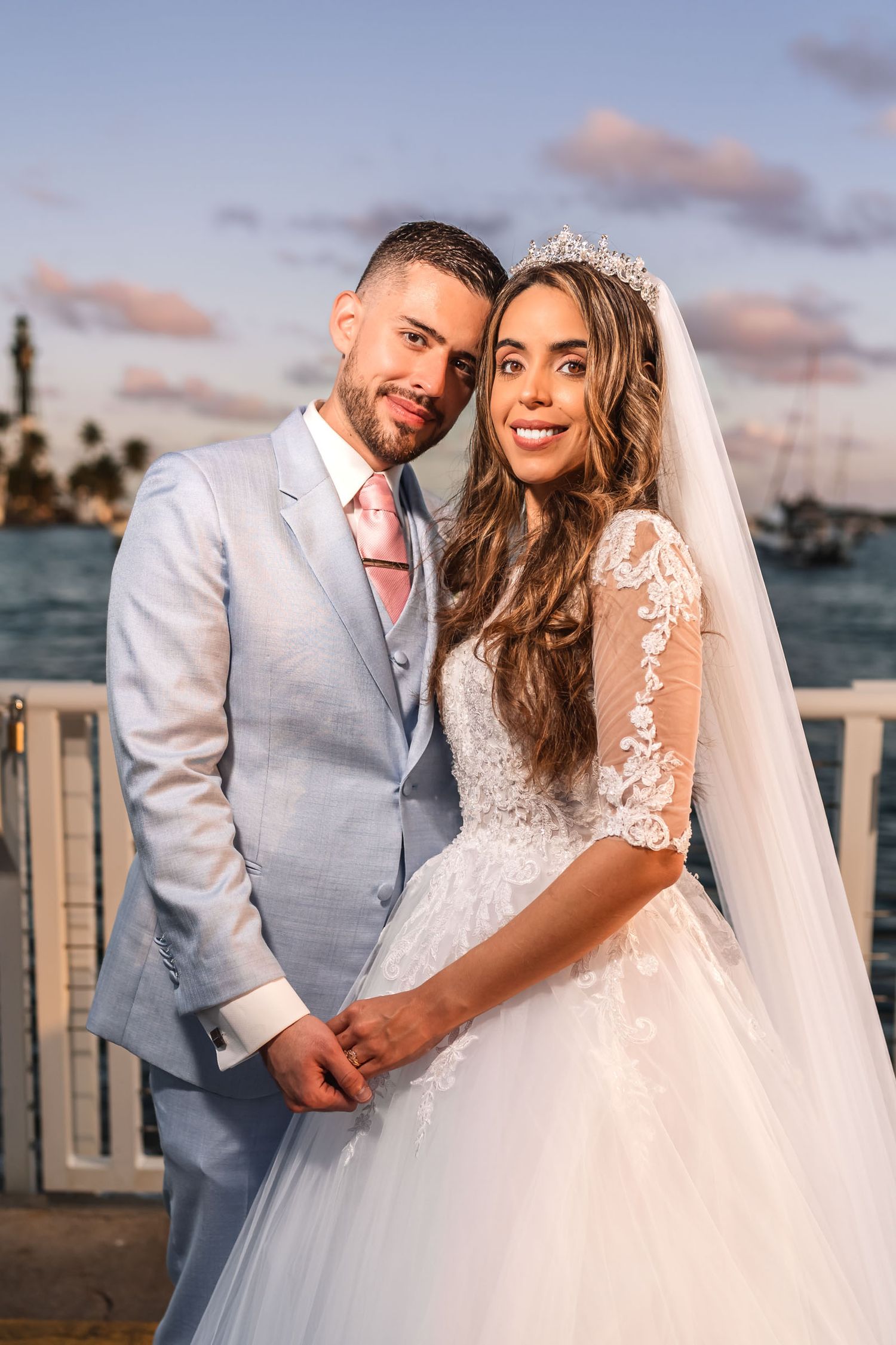 Novios sonrientes tomados de la mano al atardecer; él traje claro, ella vestido encaje, velo y tiara. Boda.