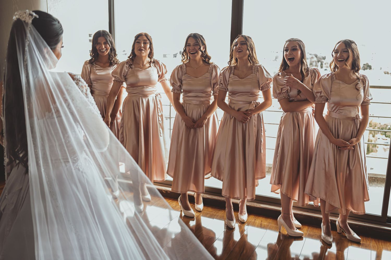 Damas de honor (vestidos rosa satinado) reaccionan felices al ver a novia (vista espalda) con velo. Boda