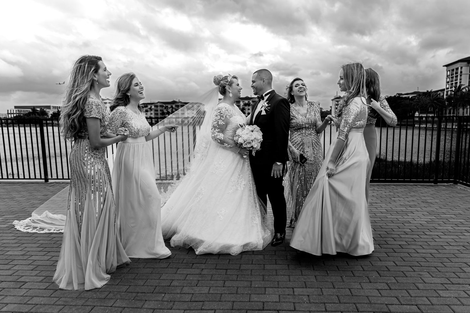 Foto B&W: Novios rodeados de damas de honor riendo en terraza; momento divertido boda en Miami al aire libre.