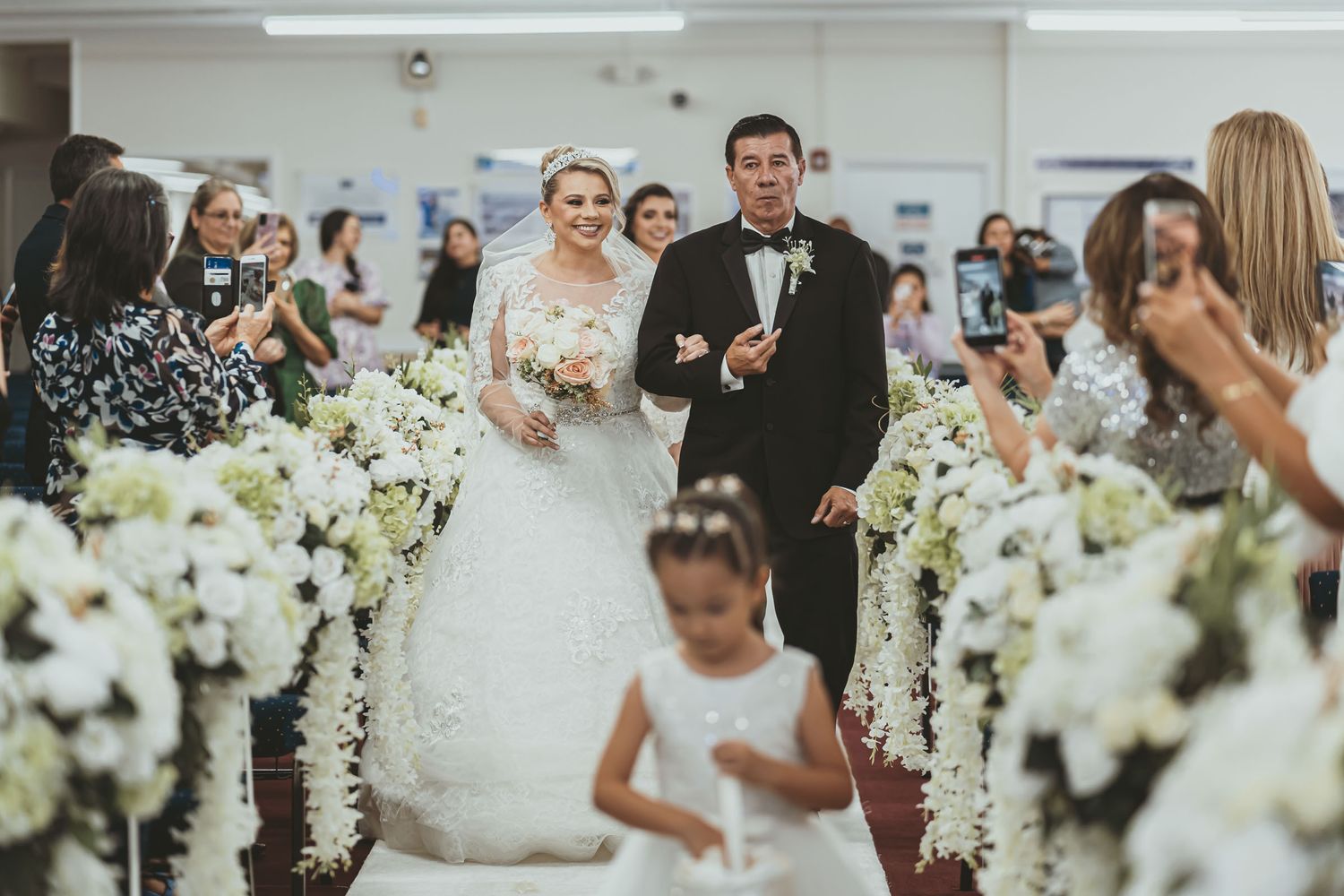 Entrada nupcial: Novia camina al altar del brazo de su padre; niña flores delante, invitados miran.