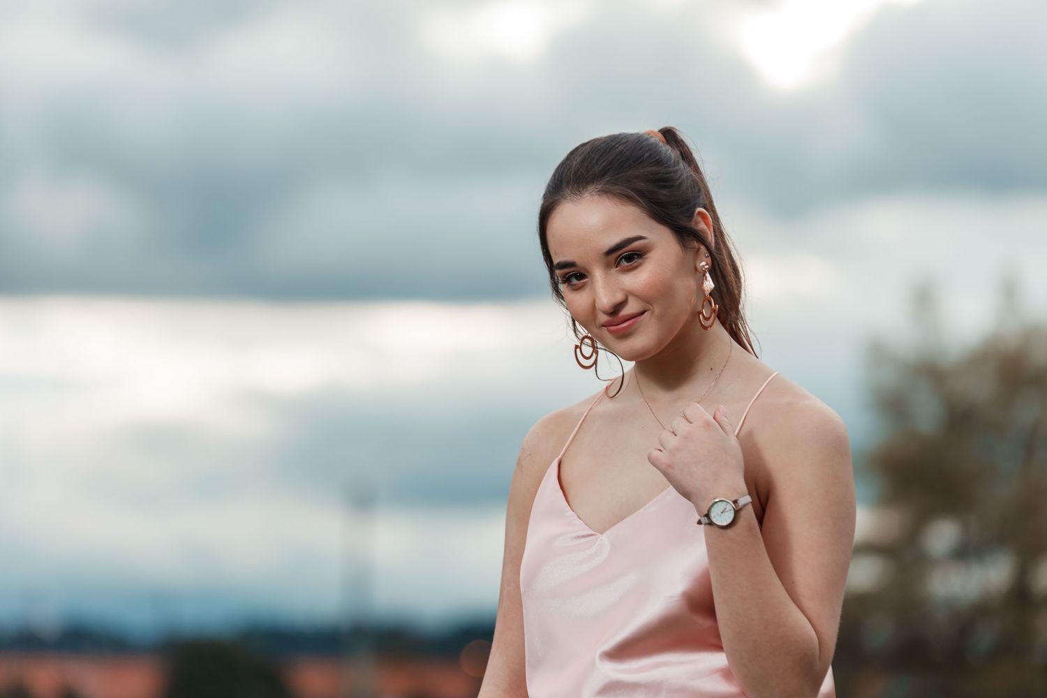 Joven mujer con top rosa y cola de caballo posa sonriente al aire libre; cielo nublado fondo. Sesión quinceañera