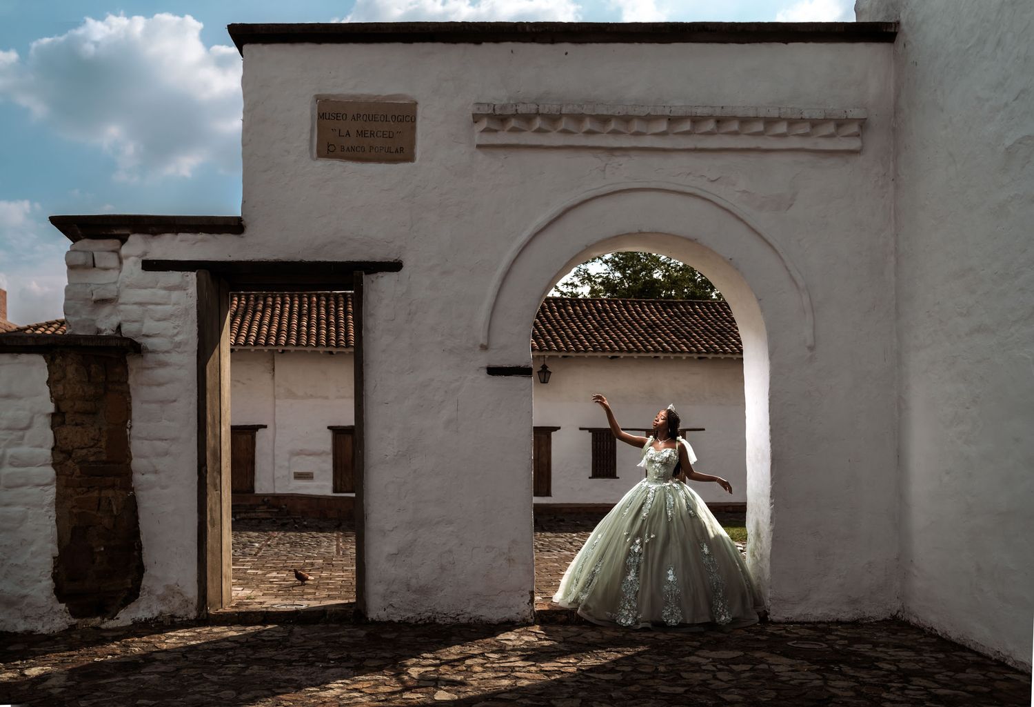 Quinceañera con vestido princesa verde/plata posa en arco de edificio colonial blanco; luz solar y sombras.