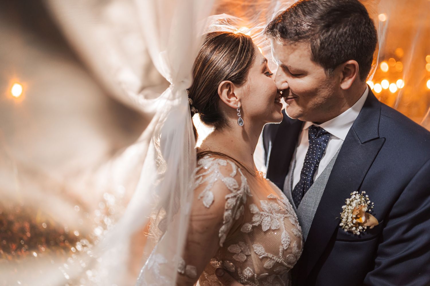 Íntima boda: pareja a punto de besarse bajo suave luz cálida y bokeh dorado. Novia con vestido de encaje y velo, novio con traje azul