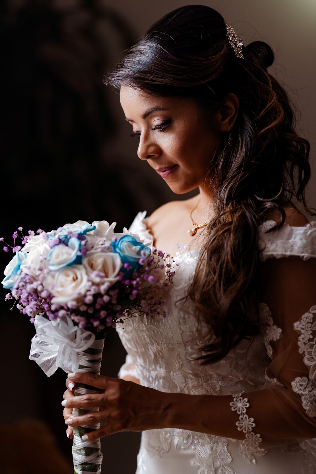 Novia mirando su ramo de rosas blancas, azules y lavanda. Viste elegante vestido blanco con detalles de encaje. Luz suave y ambiente íntimo.