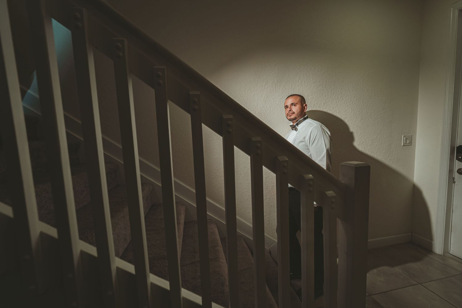 Novio con camisa y pajarita mira desde escalera con luz dramática; preparación boda interior.