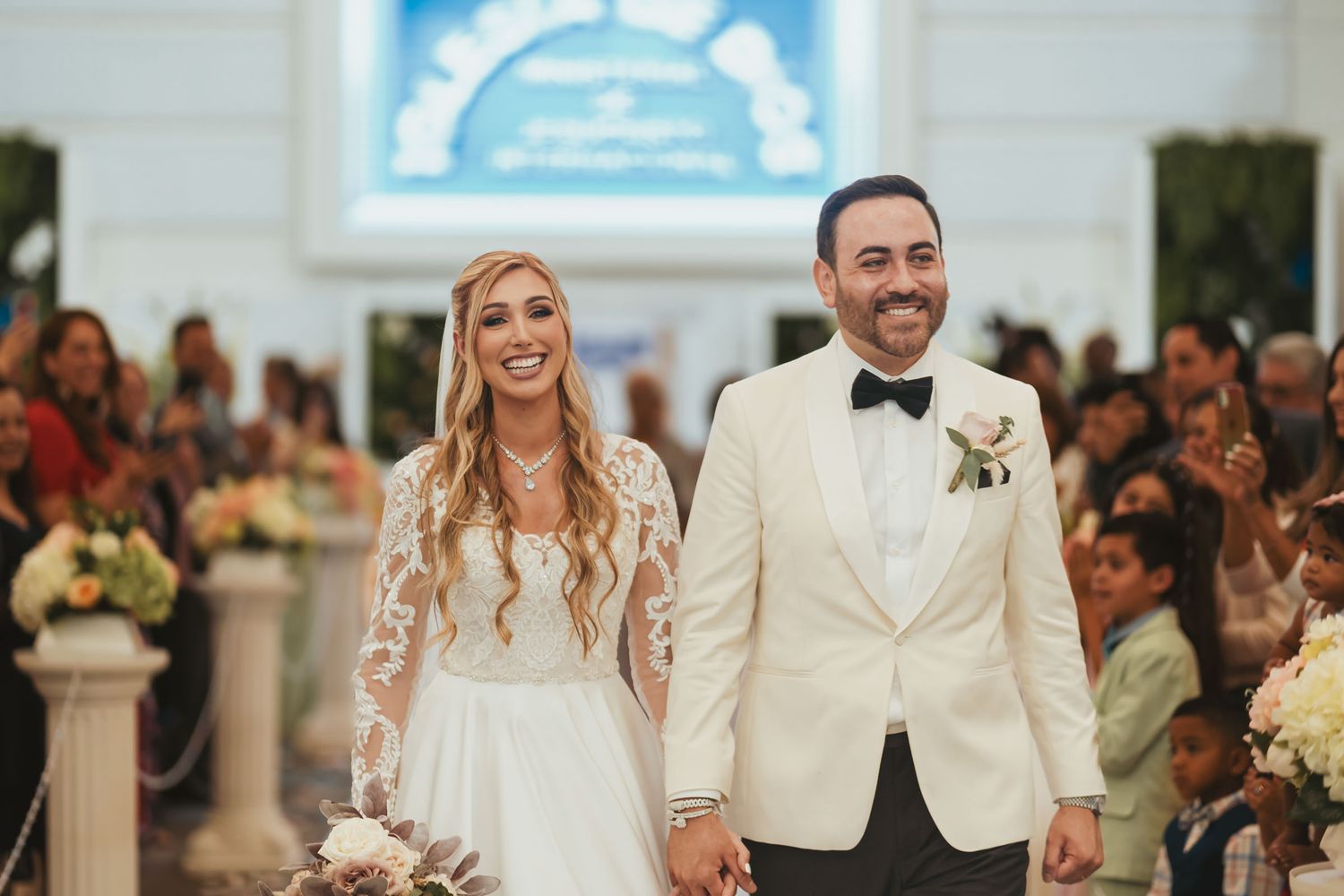 Novios caminan sonrientes por pasillo de iglesia con decoración floral. Boda IDMJI en Weston, Florida