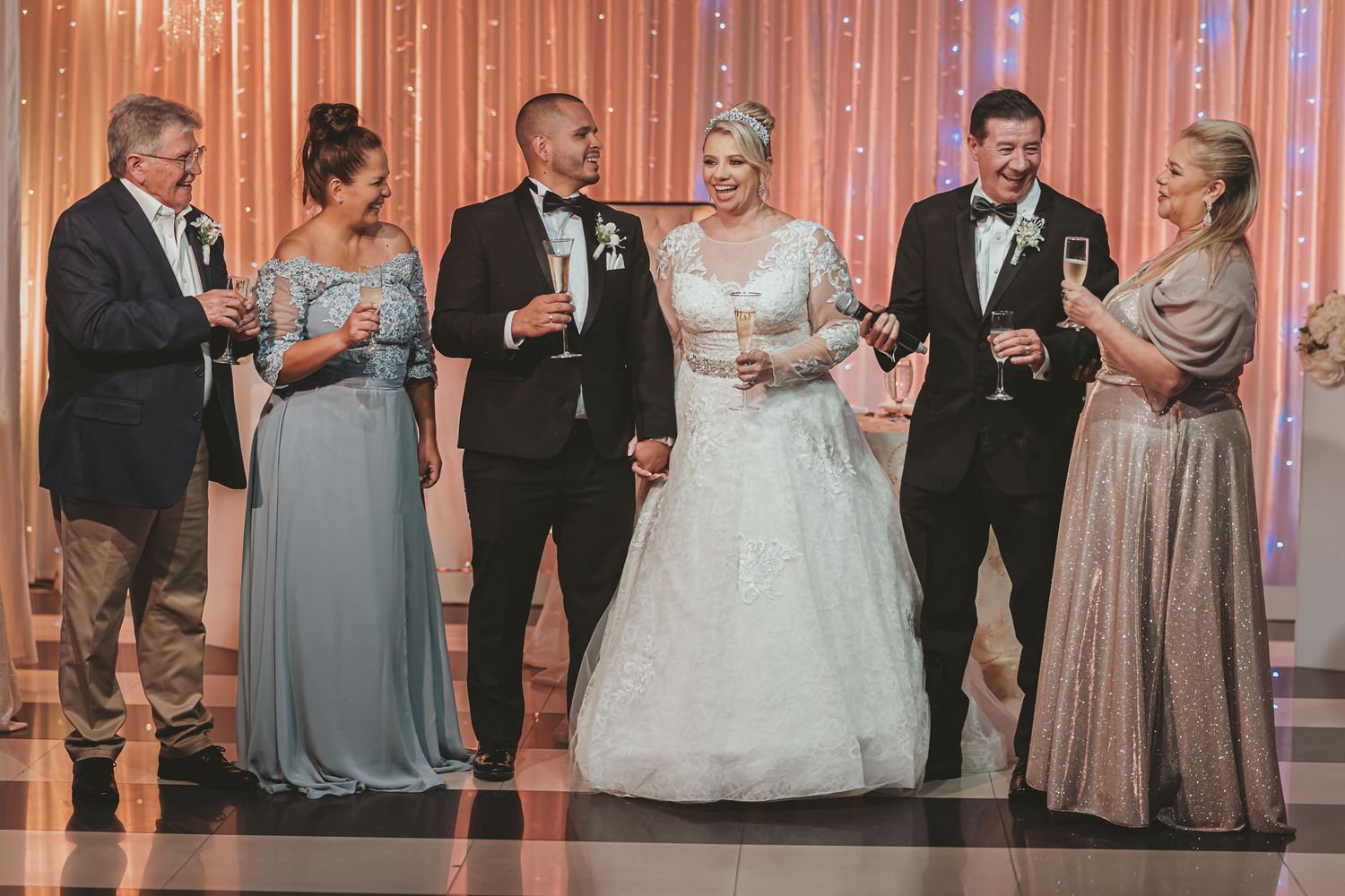 Novios sonrientes brindan con sus padres o familiares sosteniendo copas de champagne en recepción de boda.