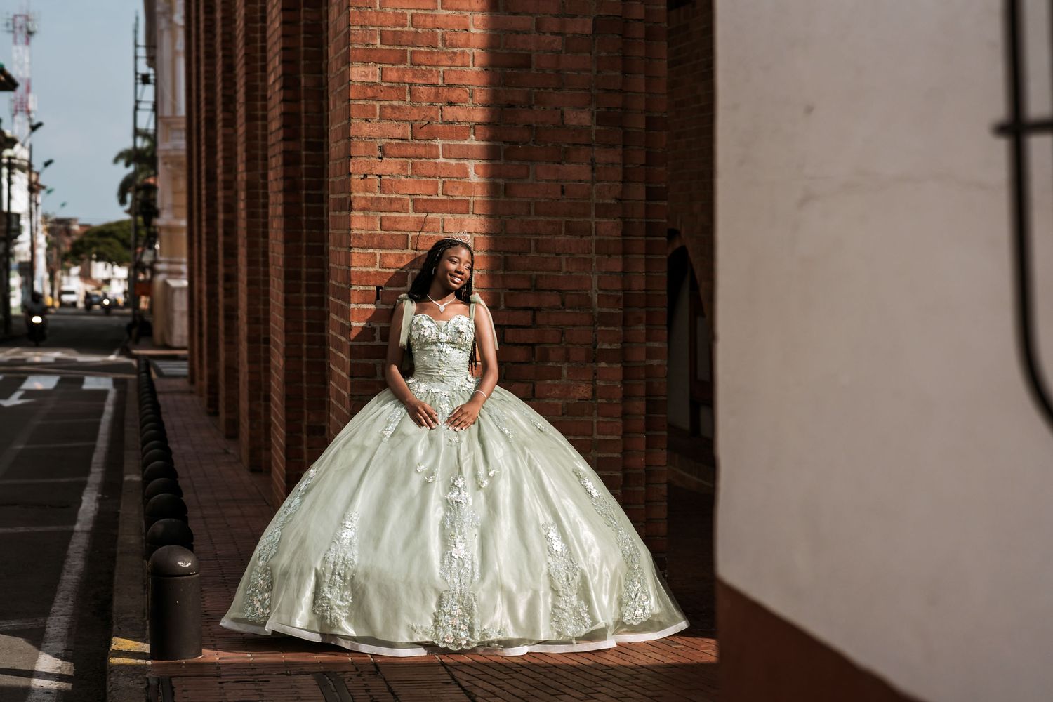 Quinceañera sonriente con vestido princesa verde/plata y tiara posa en exterior junto a pared ladrillo; luz. Sesión de 15