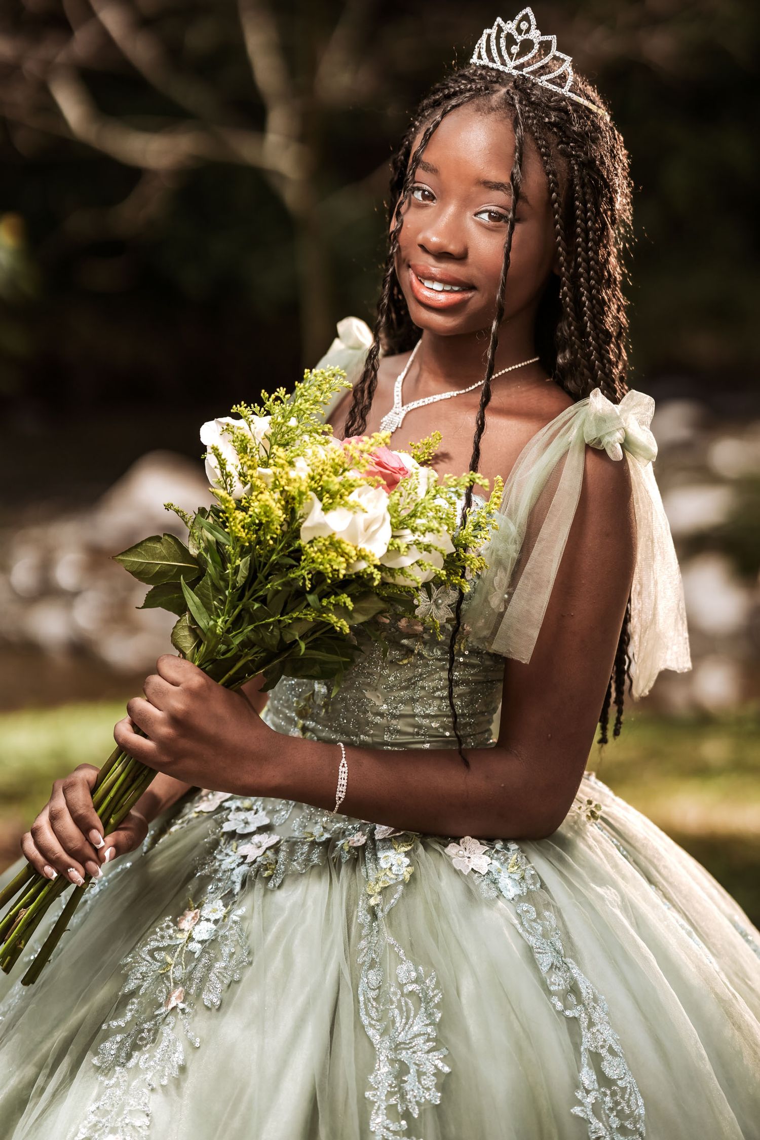 Retrato quinceañera afrodescendiente (trenzas, vestido verde, tiara) sostiene ramo flores en parque.