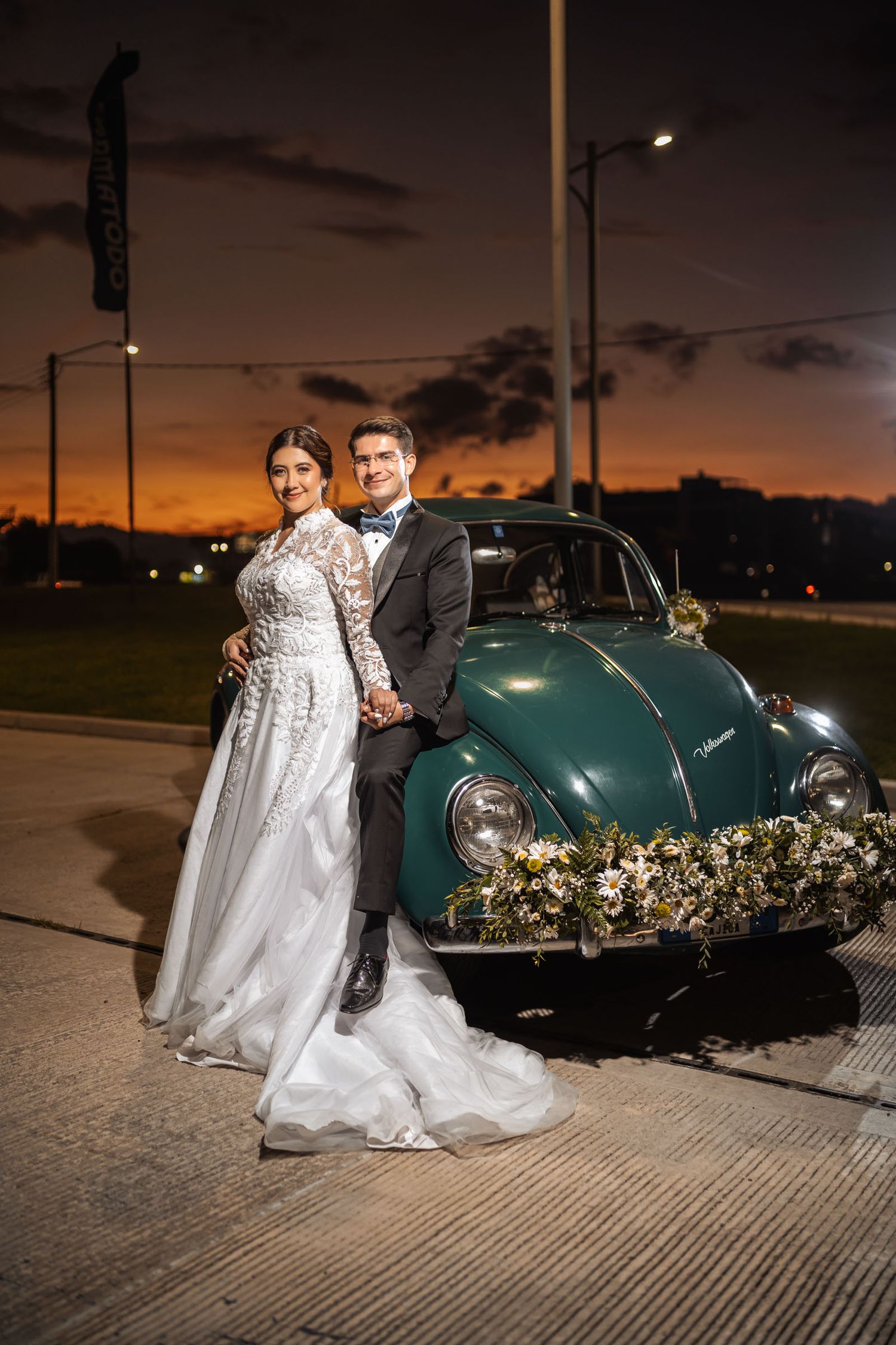 Novios sonríen posando junto a coche verde clásico al anochecer, con luces de ciudad al fondo. Boda exterior.