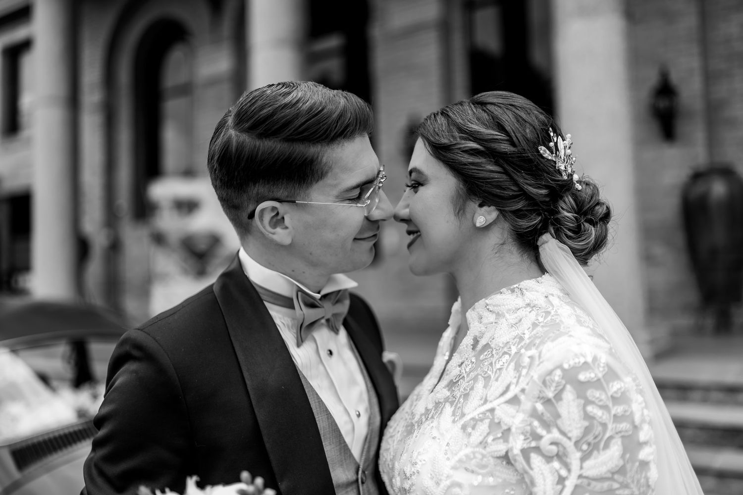 Novios se miran nariz con nariz; retrato íntimo de boda en blanco y negro frente a edificio. Él esmoquin, ella vestido encaje.