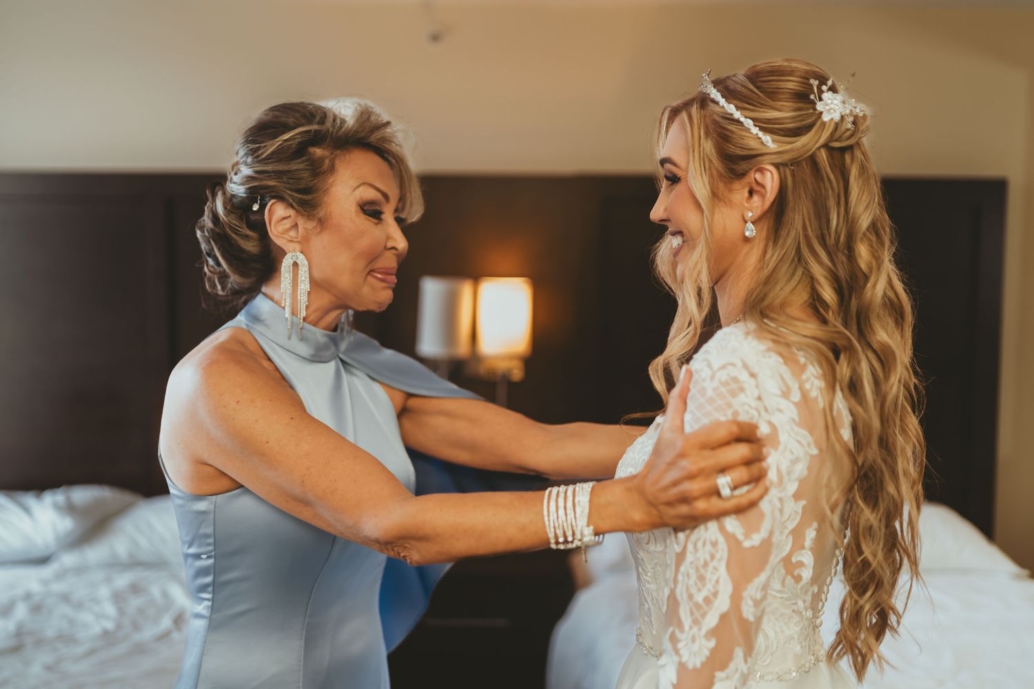 Madre (vestido azul) ayuda a novia (vestido encaje, tocado perlas) a prepararse en habitación. Boda.