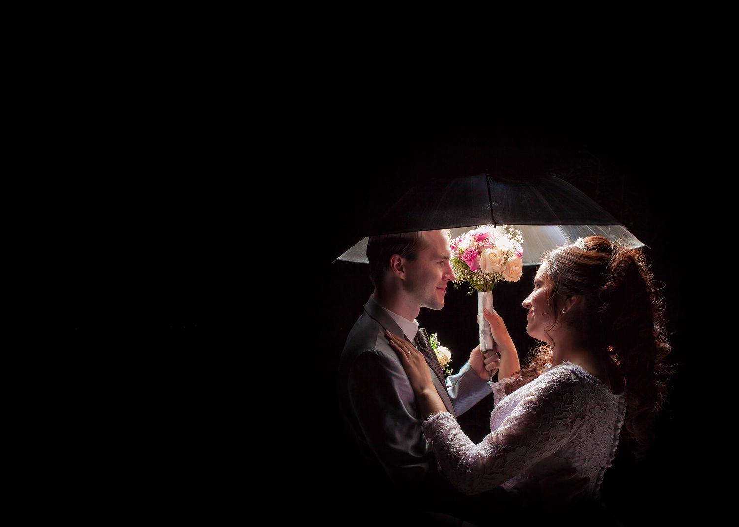 Novios se miran bajo paraguas iluminado sosteniendo ramo; fondo oscuro. Foto boda creativa íntima.