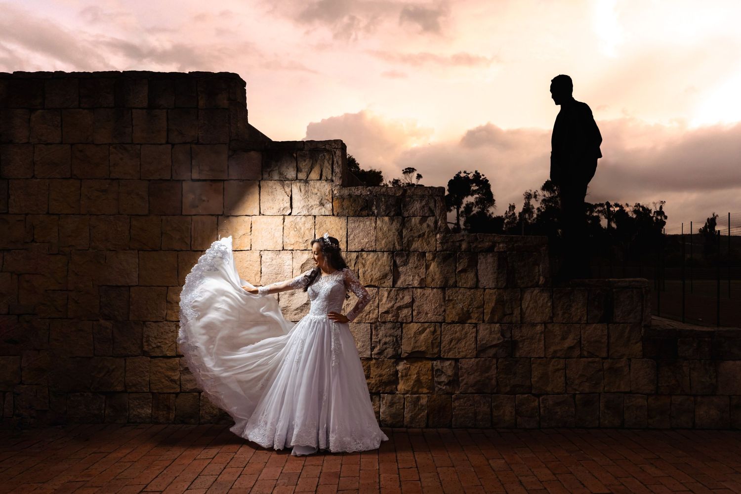 Novia con vestido blanco ondeante y corona mirando hacia silueta de novio en ruinas de ladrillo al atardecer. Ambiente contemplativo