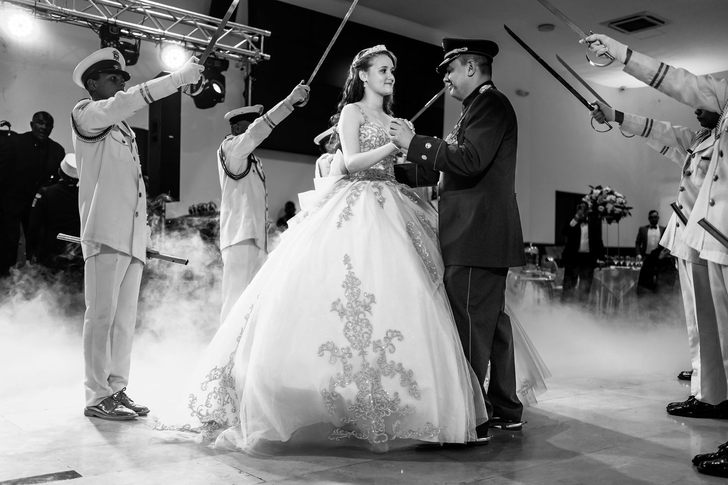Foto B&W: Quinceañera baila con padre/escolta bajo arco de sables de cadetes; niebla pista. Fiesta 15.