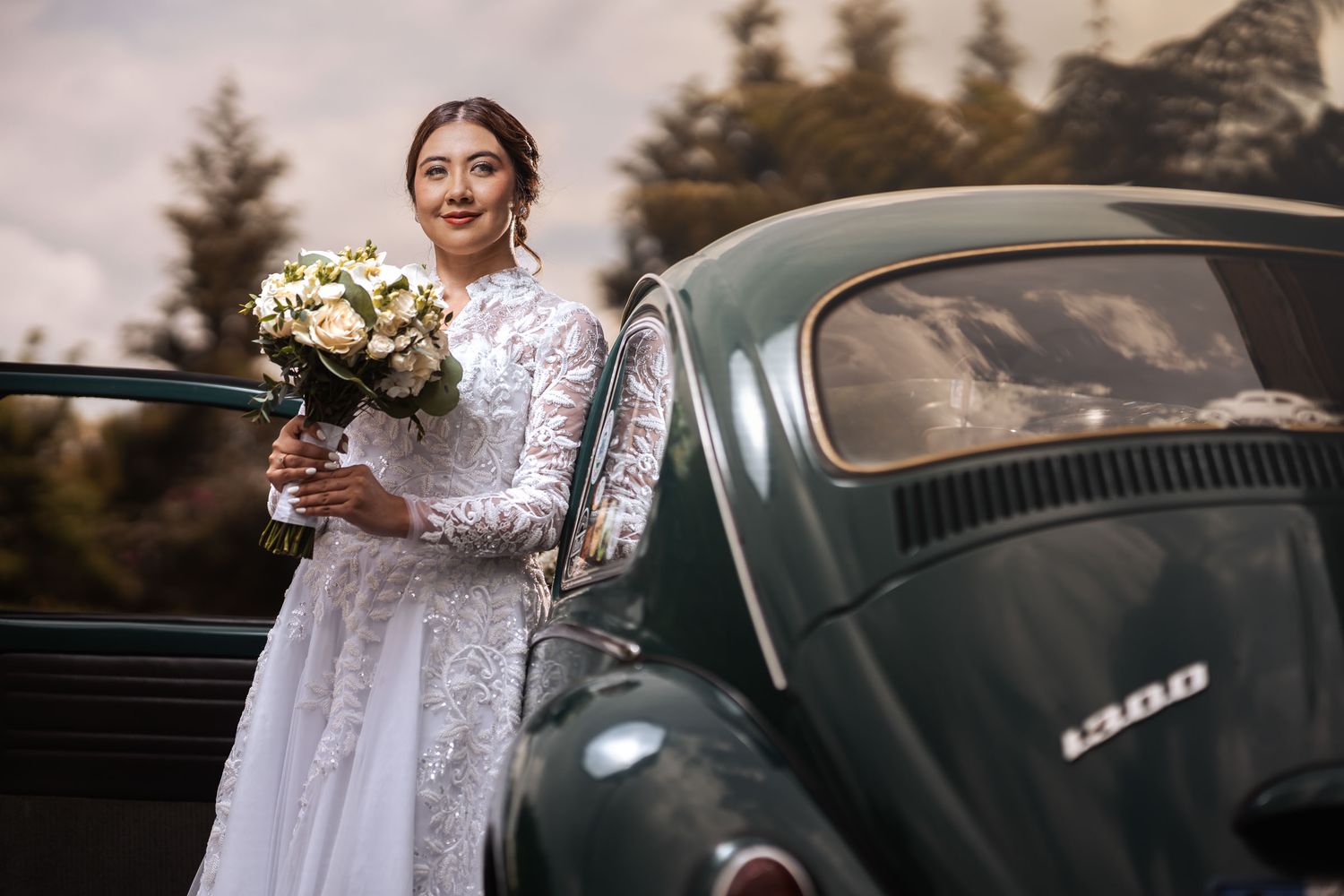 Novia con vestido de encaje y ramo sonríe apoyada en coche clásico VW Escarabajo verde en exterior. Fotografía boda.