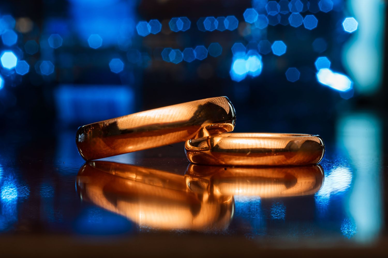 Detalle macro: dos anillos de boda dorados reflejados sobre superficie con fondo de luces bokeh azules brillantes. Boda