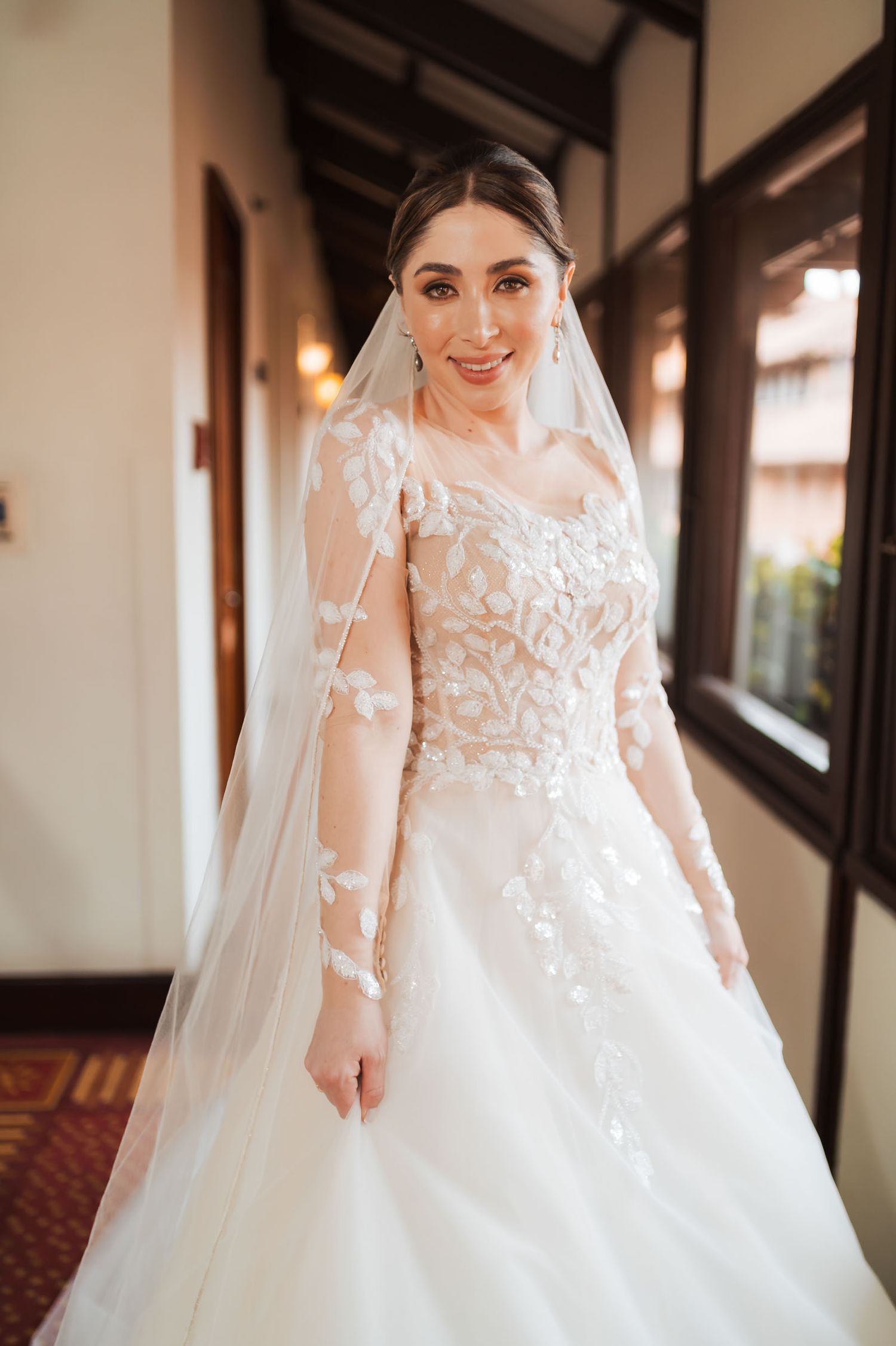 Novia sonriente posa en pasillo; luce vestido de novia bordado manga larga, falda tul y velo largo. Boda.