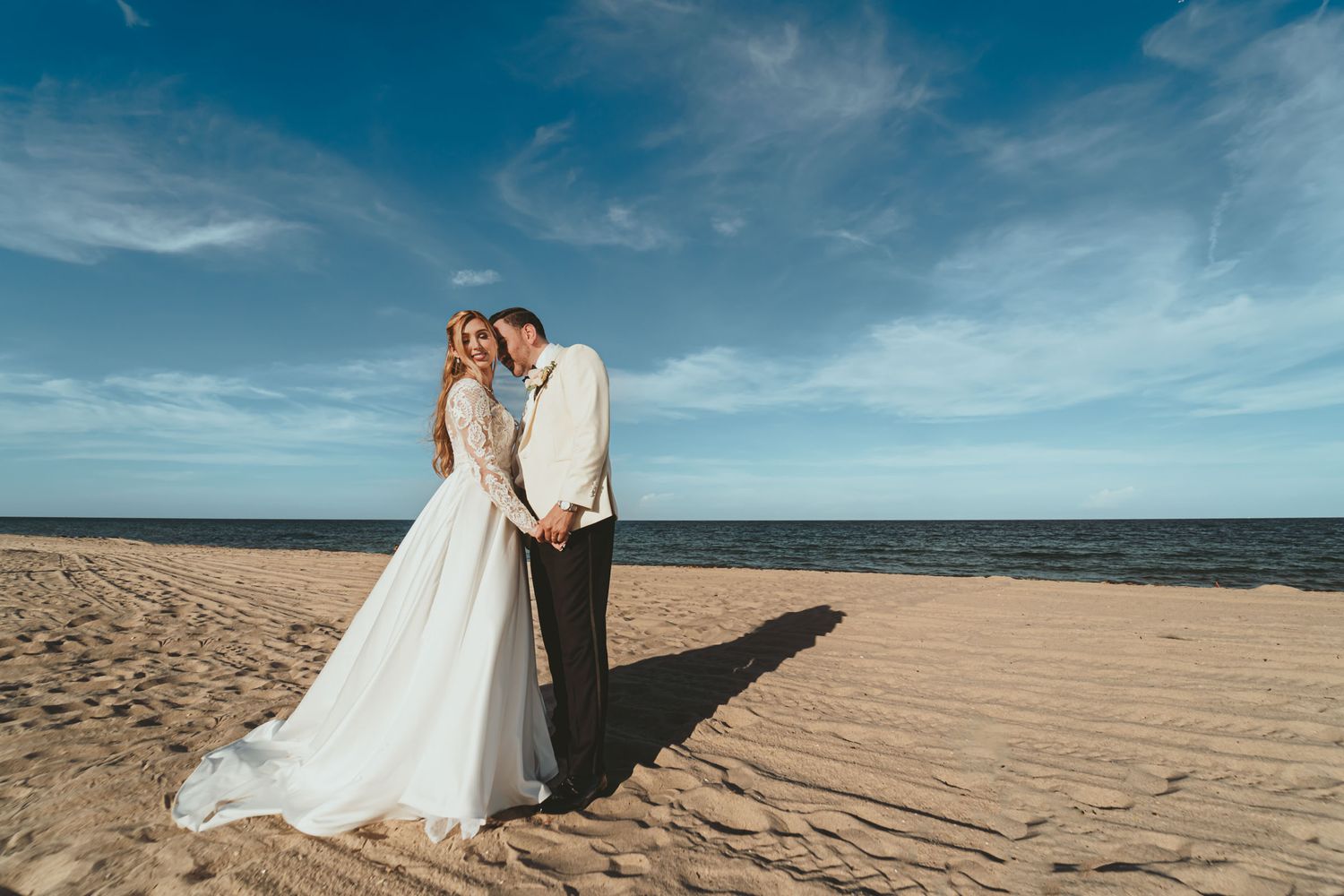 Novios se besan en playa; él chaqueta clara, ella vestido blanco. Cielo azul, mar fondo. Boda destino.