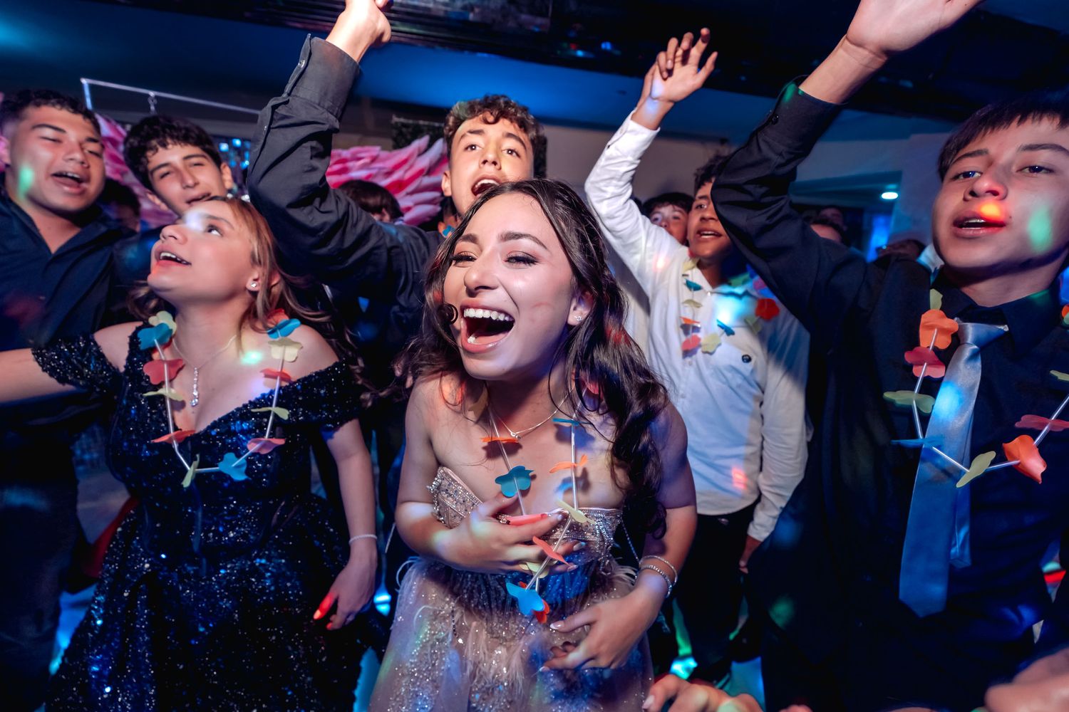 Grupo de jóvenes baila enérgicamente en pista iluminada durante fiesta de quinceañera. Brazos arriba, pura diversión.