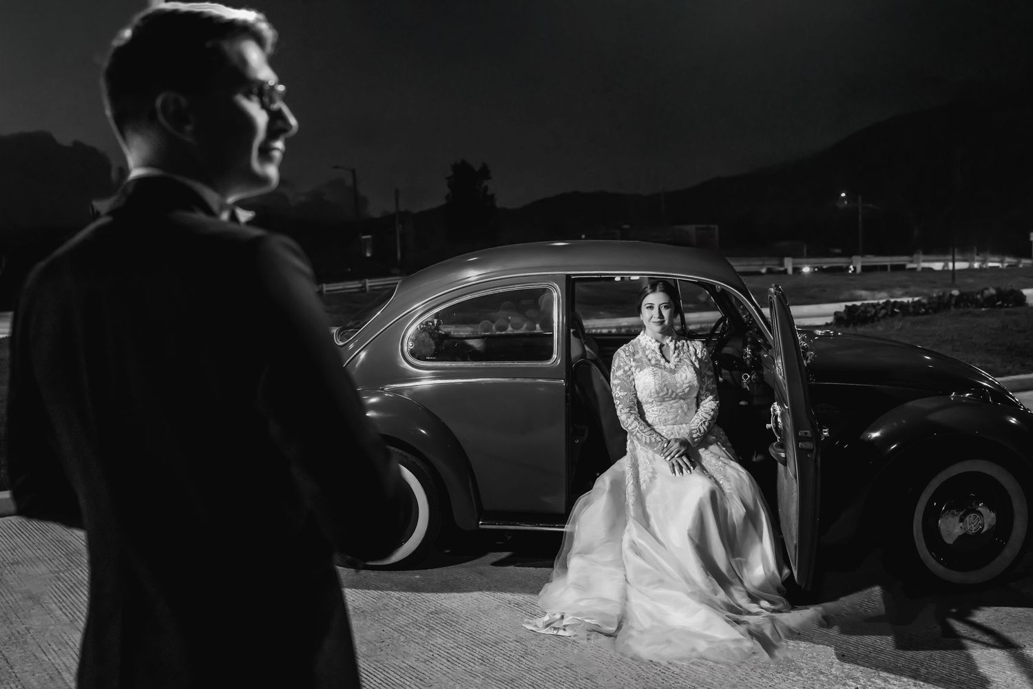 Silueta de novio mira a novia sentada en coche clásico de noche. Foto de boda blanco y negro, momento dramático.