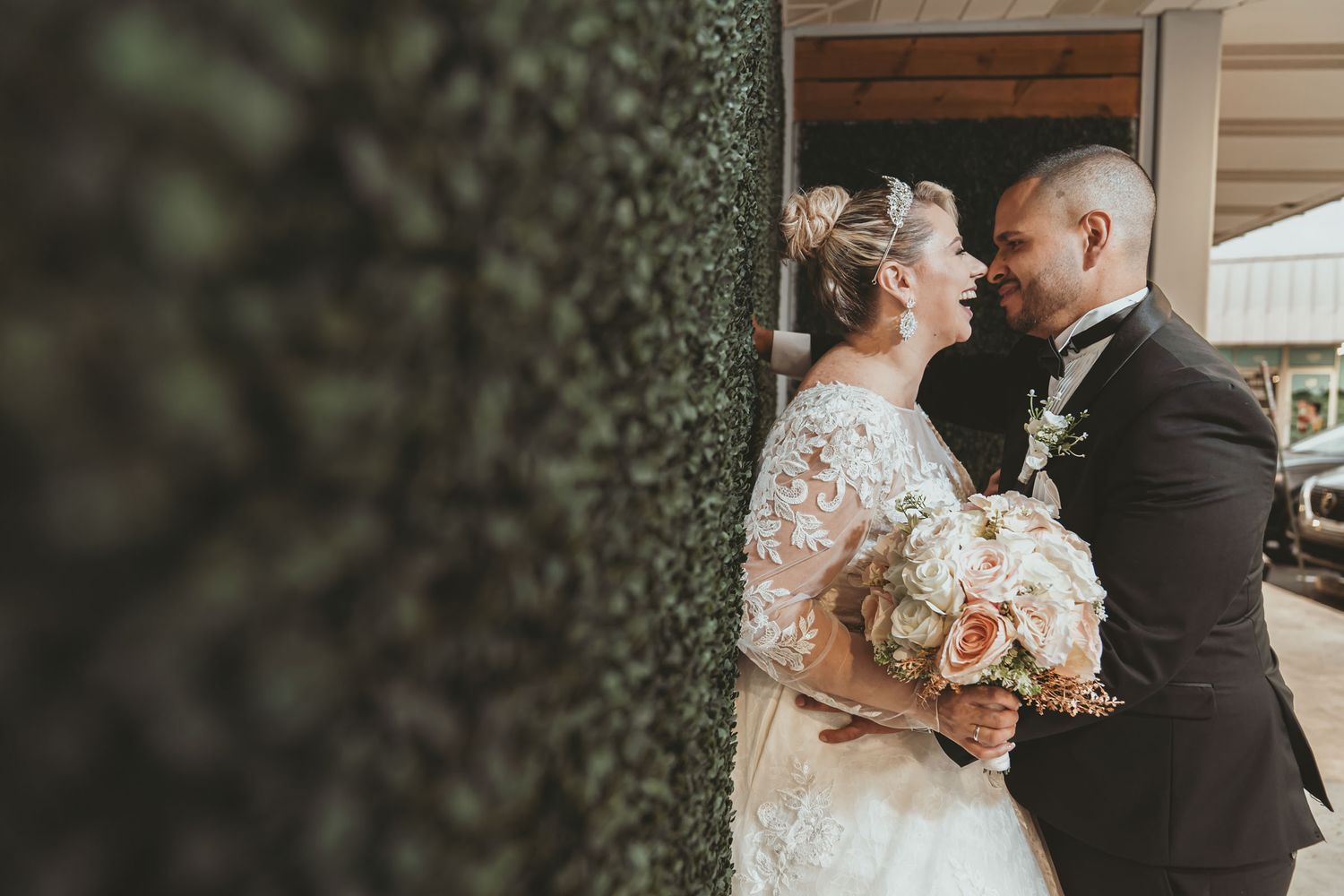 Novios se miran sonrientes y de cerca junto a pared de follaje verde; novia sostiene ramo. Boda.