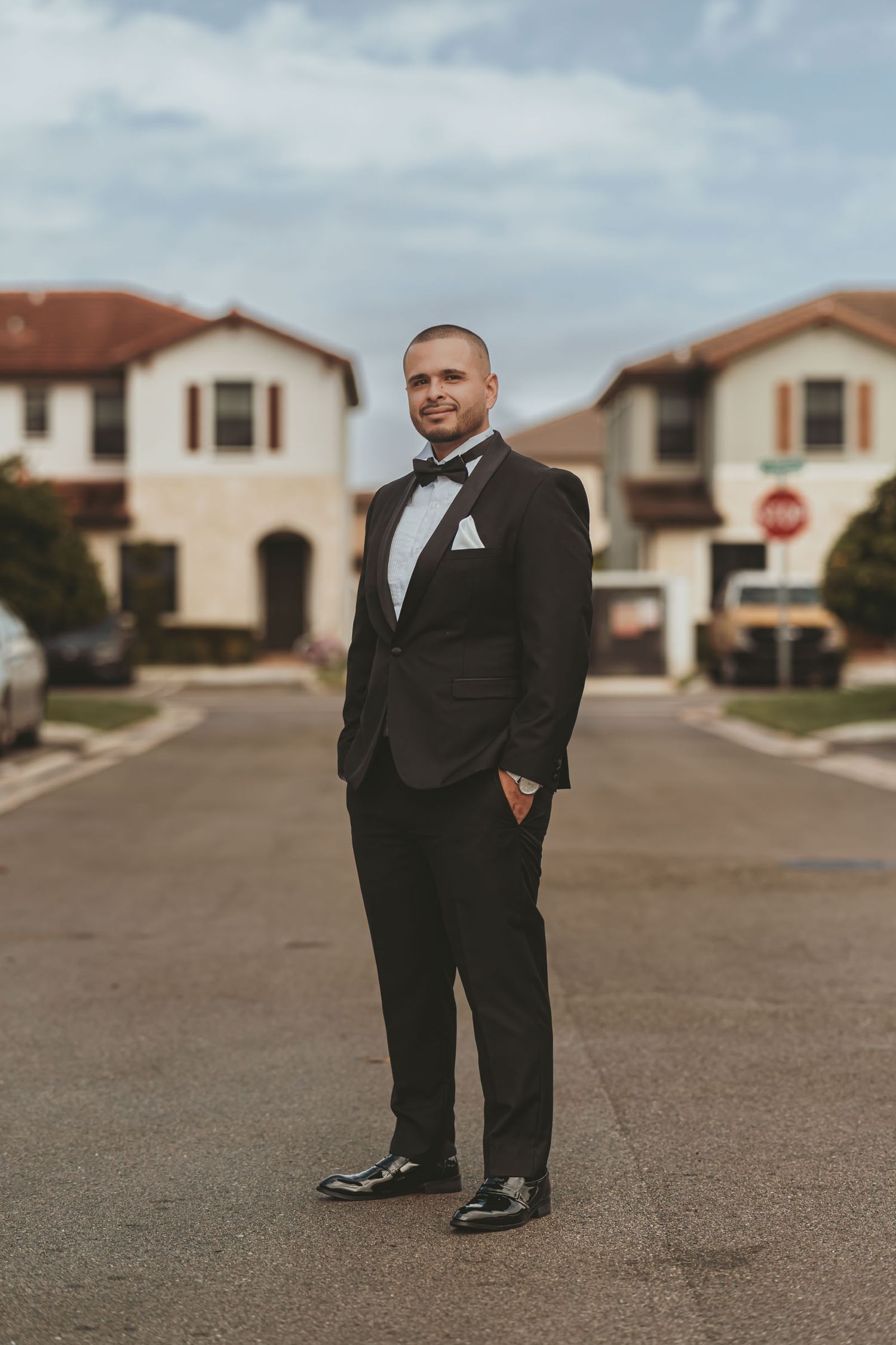 Novio elegante con esmoquin posa en medio de calle residencial bajo cielo nublado; retrato boda exterior en Miamia.