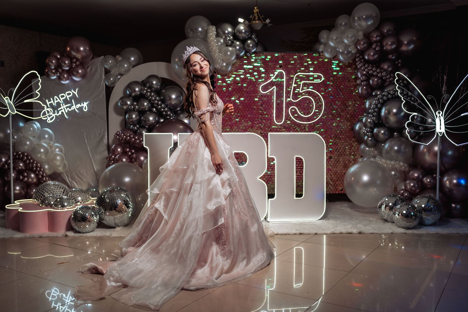 Quinceañera celebra sus 15 años; posa mirando sobre el hombro con vestido rosa y tiara ante decoración festiva.
