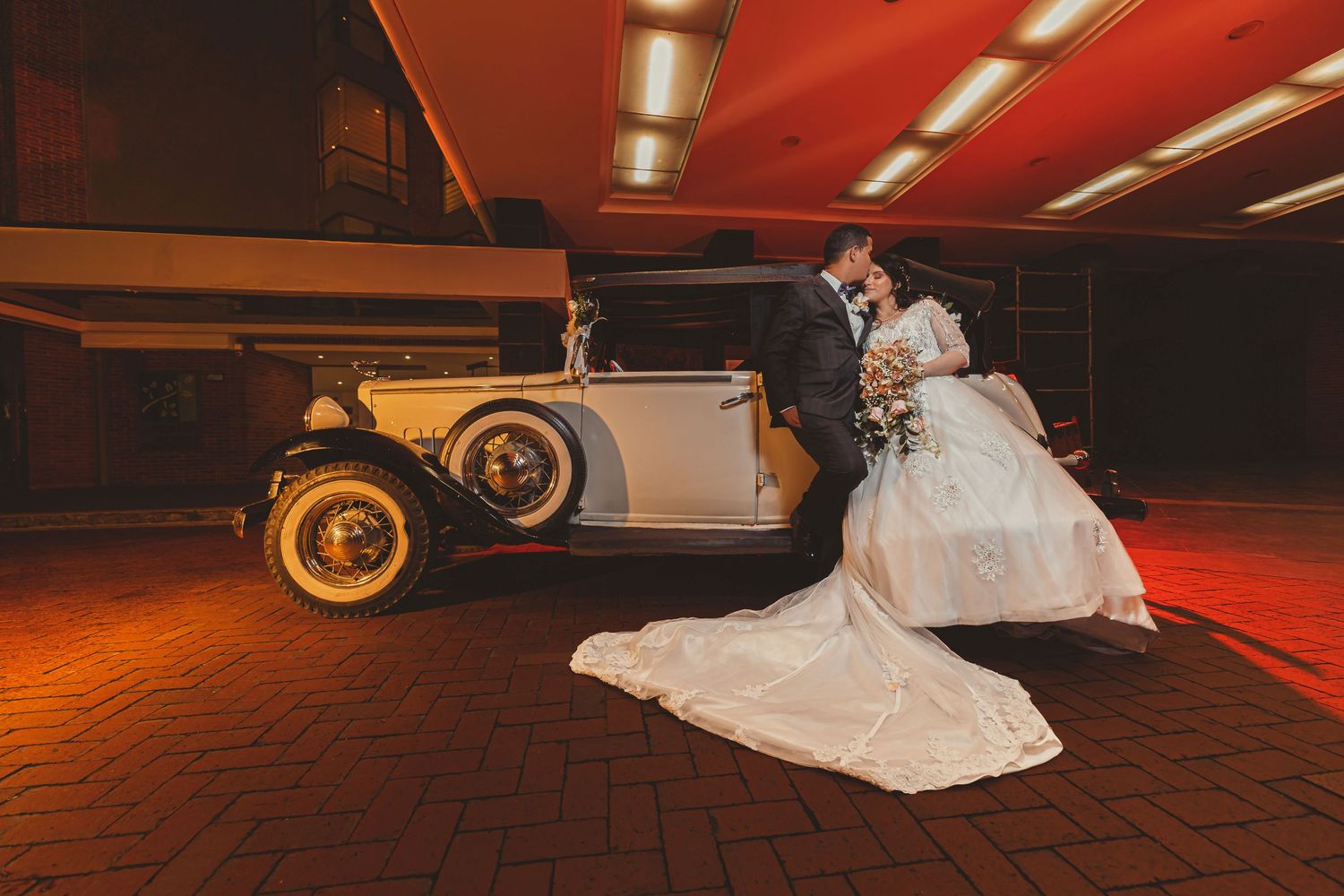 Novios se besan junto a coche clásico convertible blanco bajo luces rojas; entrada edificio. Boda elegante.