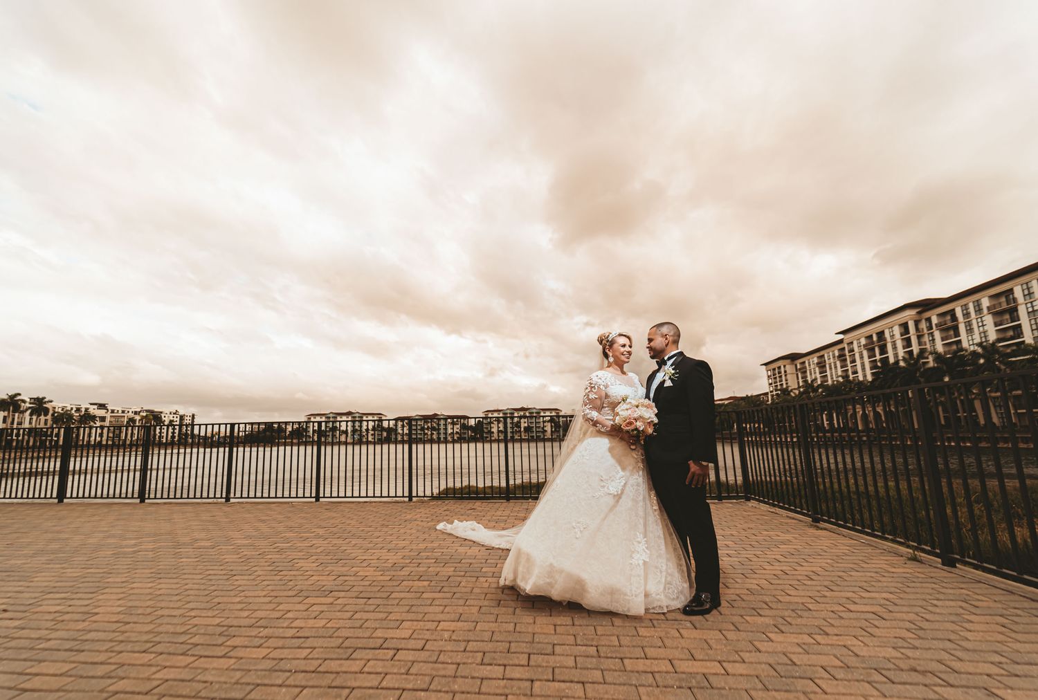 Novios posan en terraza exterior junto a lago bajo cielo nublado de Miami; vista amplia. Fotografía boda.