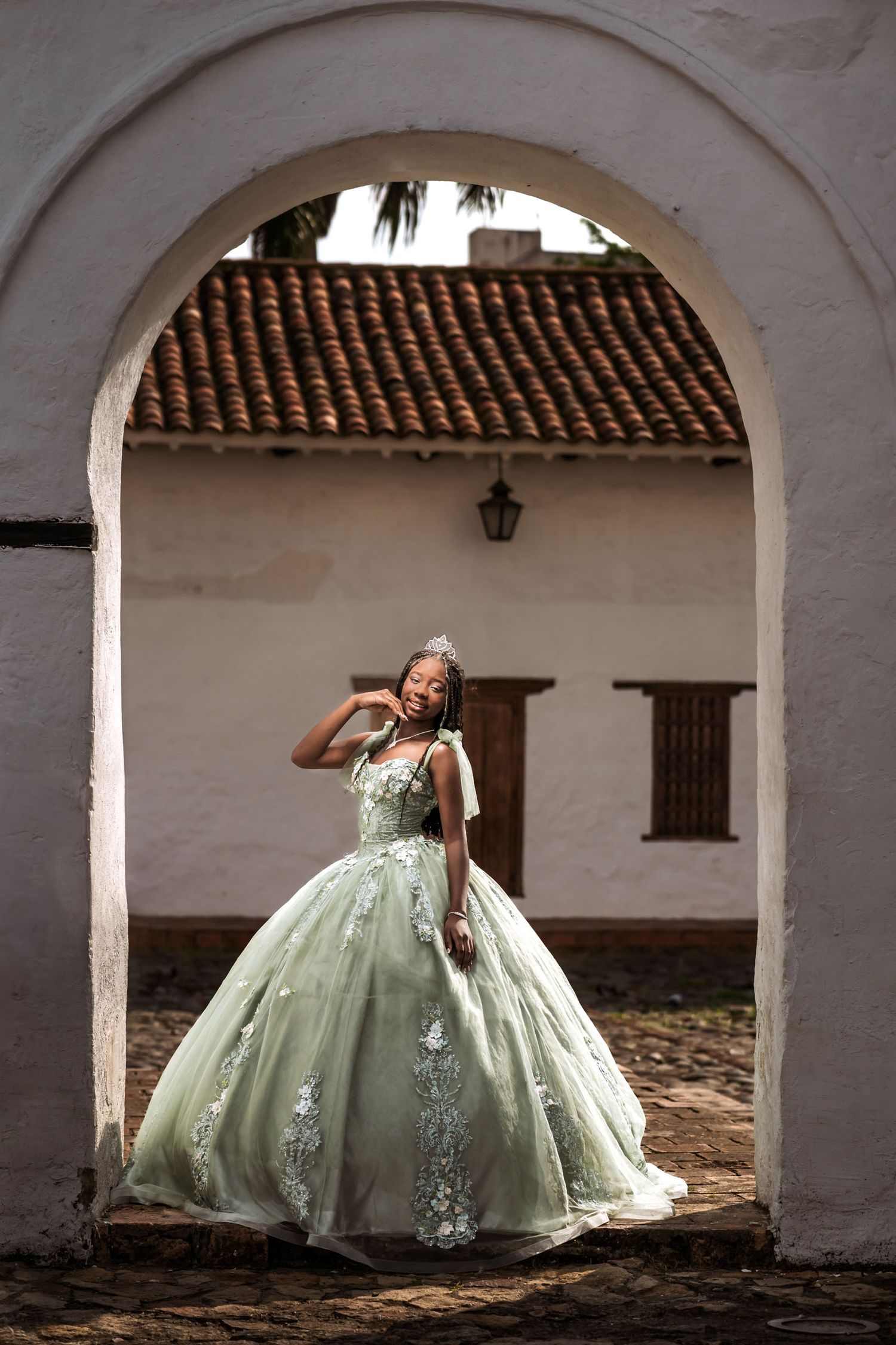 Quinceañera con vestido verde/plata y tiara posa elegante bajo arco edificio colonial blanco; sesión fotos.