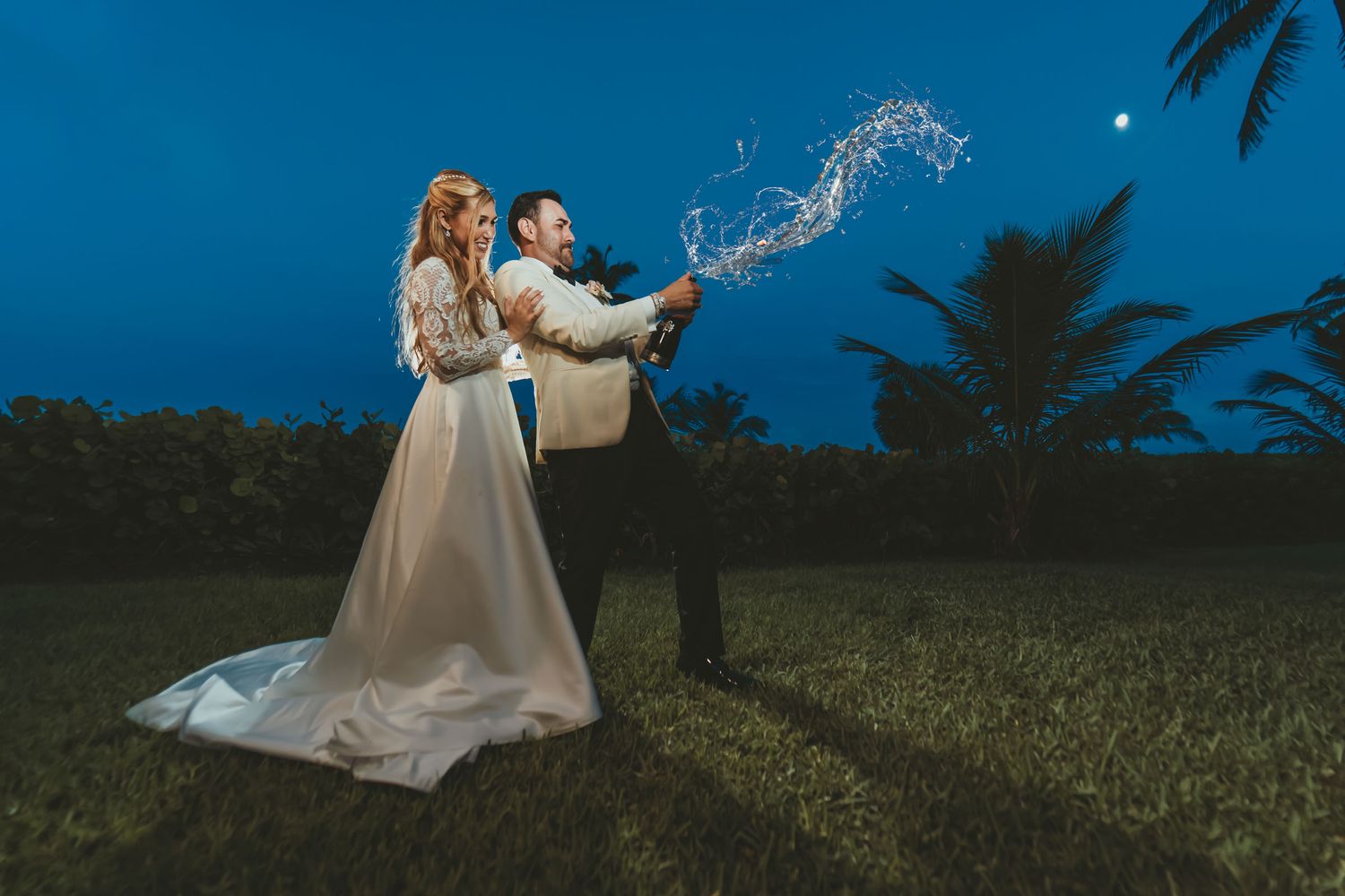 Novios celebran de noche en exterior: novio destapa botella de champagne con chorro espumoso; novia sonríe. Boda Miami