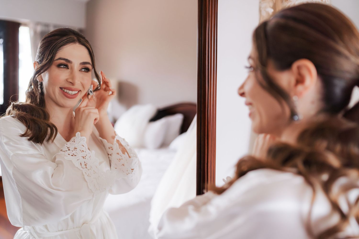 Novia sonriente con bata blanca se ajusta el arete mirándose en espejo; preparación boda en dormitorio.