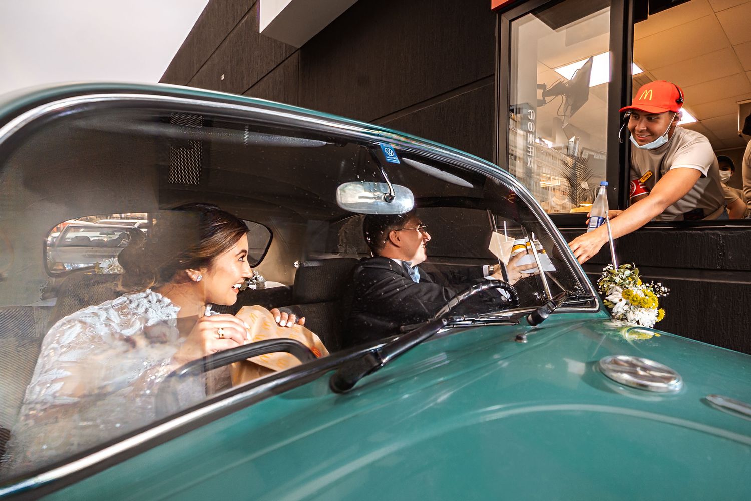 Novios sonríen juntos sentados dentro de coche clásico verde mientras compran en Mc Donalds. Foto de boda íntima.