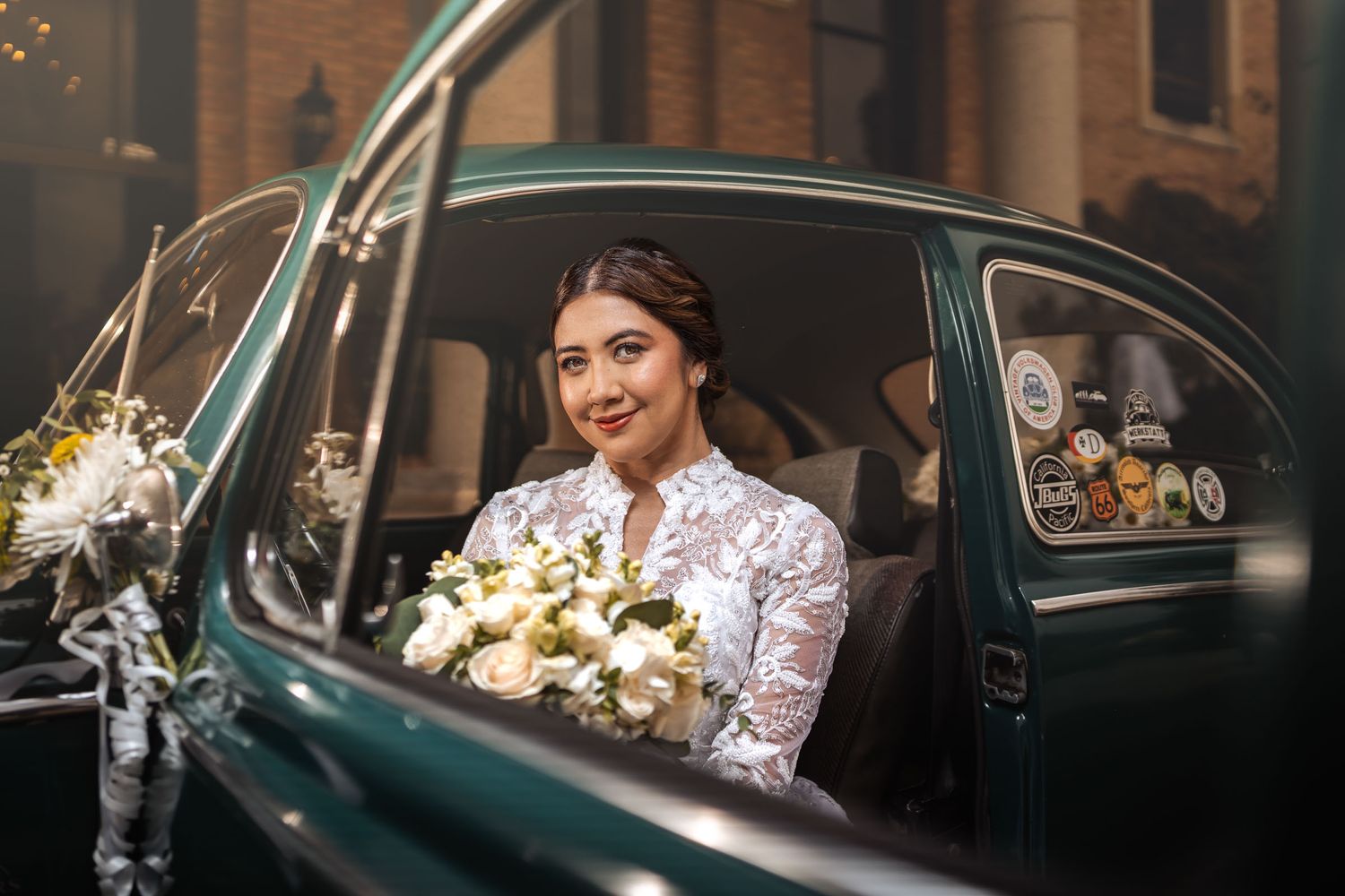 Novia sonriente con vestido de encaje y ramo sentada en coche clásico VW Escarabajo verde; puerta abierta. Fotografía de boda.