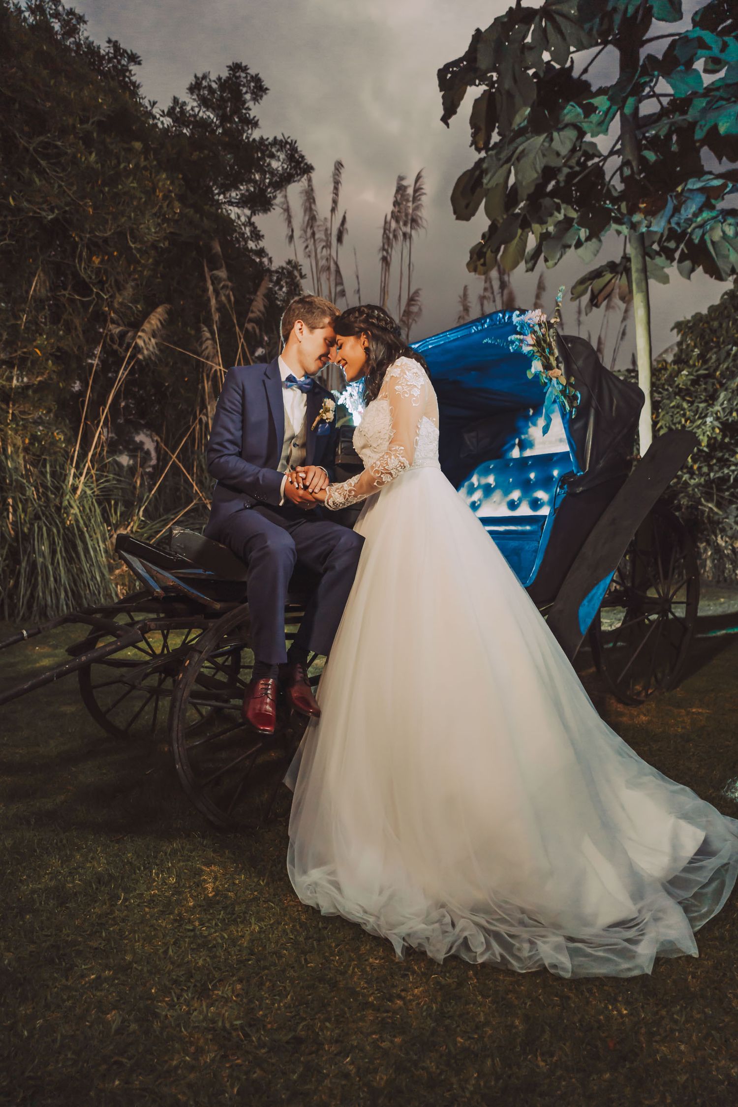 Novios posando de noche en un jardín junto a una carruaje vintage en Cajicá.