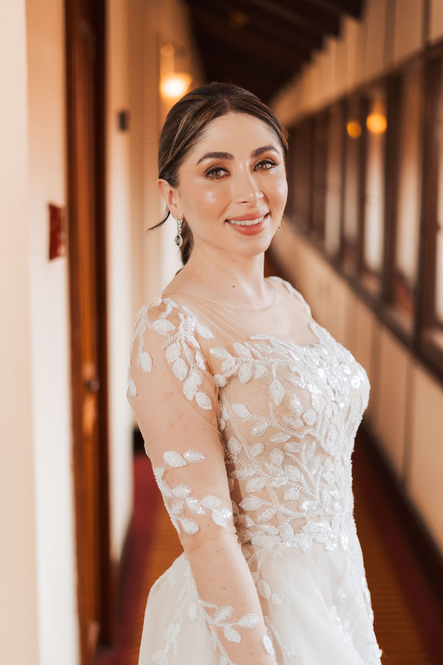 Retrato de novia sonriente en pasillo interior; luce vestido de novia bordado manga larga y velo. Boda.