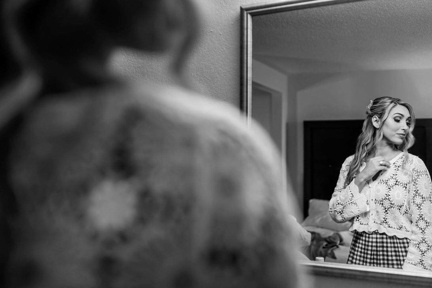 Foto B&W: Reflejo de novia sonriente (blusa encaje, falda cuadros, tocado) en espejo; preparación boda.
