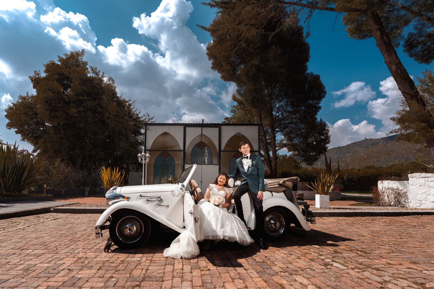 Novia sentada en coche clásico junto a novio en patio colonial