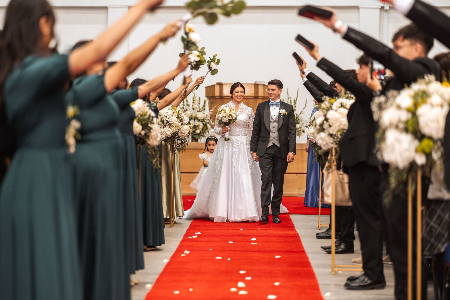 Novios caminan sonrientes por pasillo de alfombra roja, invitados celebran su paso levantando flores. Boda IDMJI.