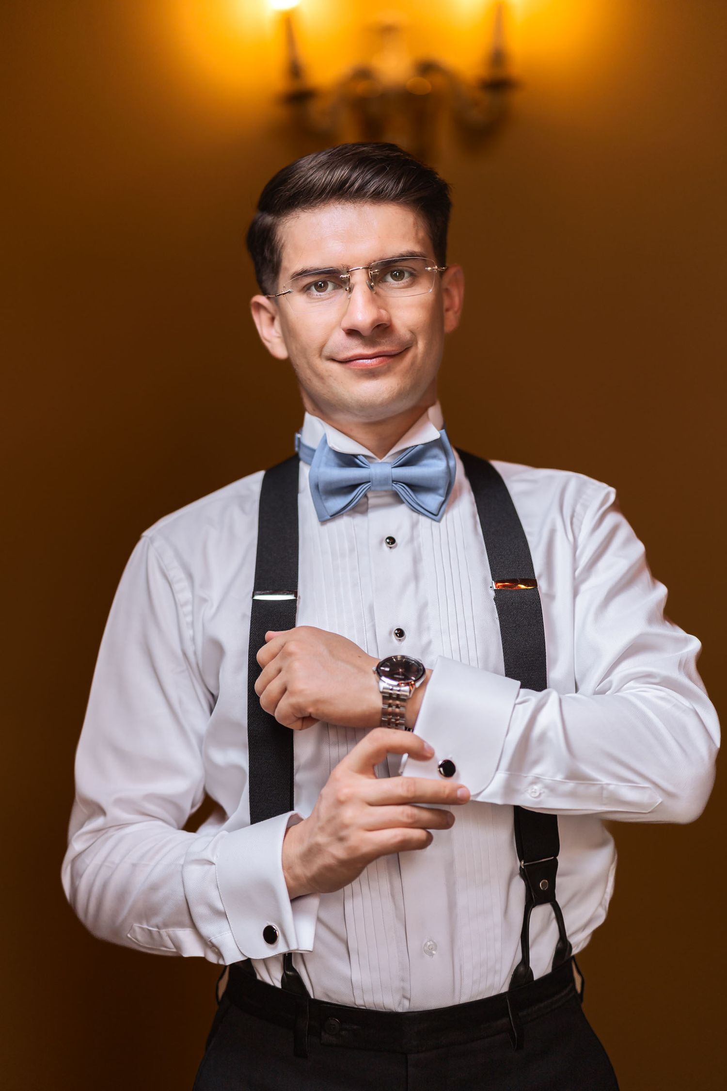 Novio con camisa, pajarita azul y tirantes ajusta su gemelo o reloj mirando a cámara; preparación antes de la boda.