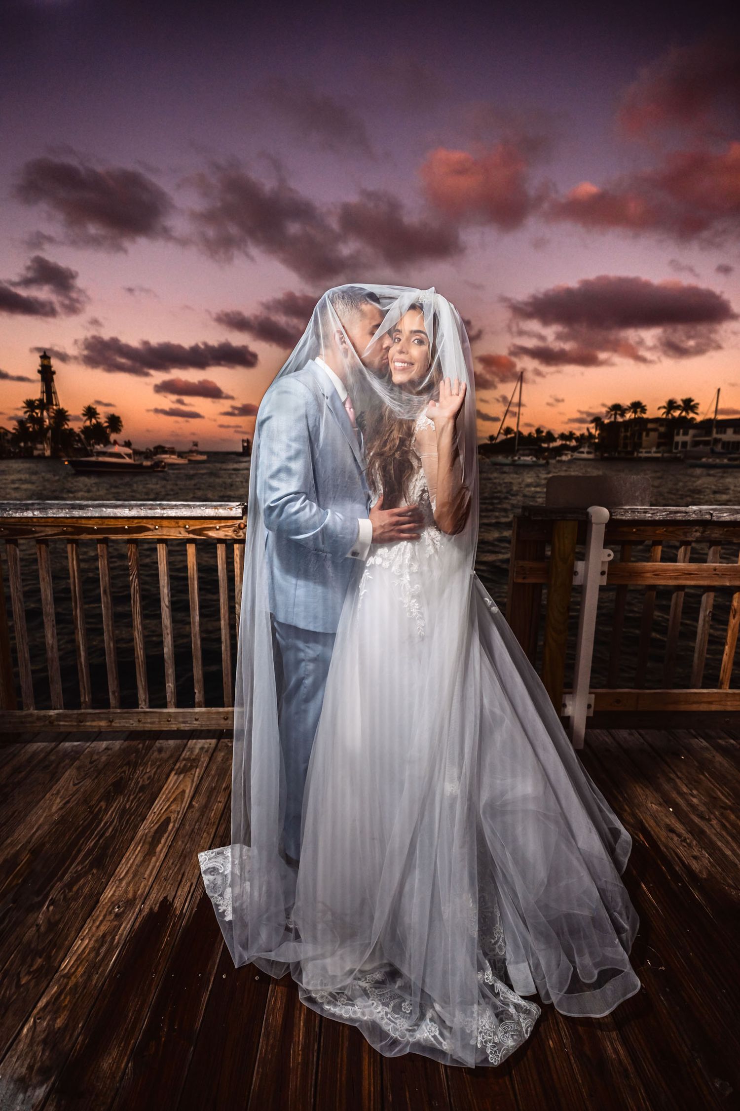 Novios abrazados bajo el velo en un muelle durante su sesión de fotos postboda al atardecer