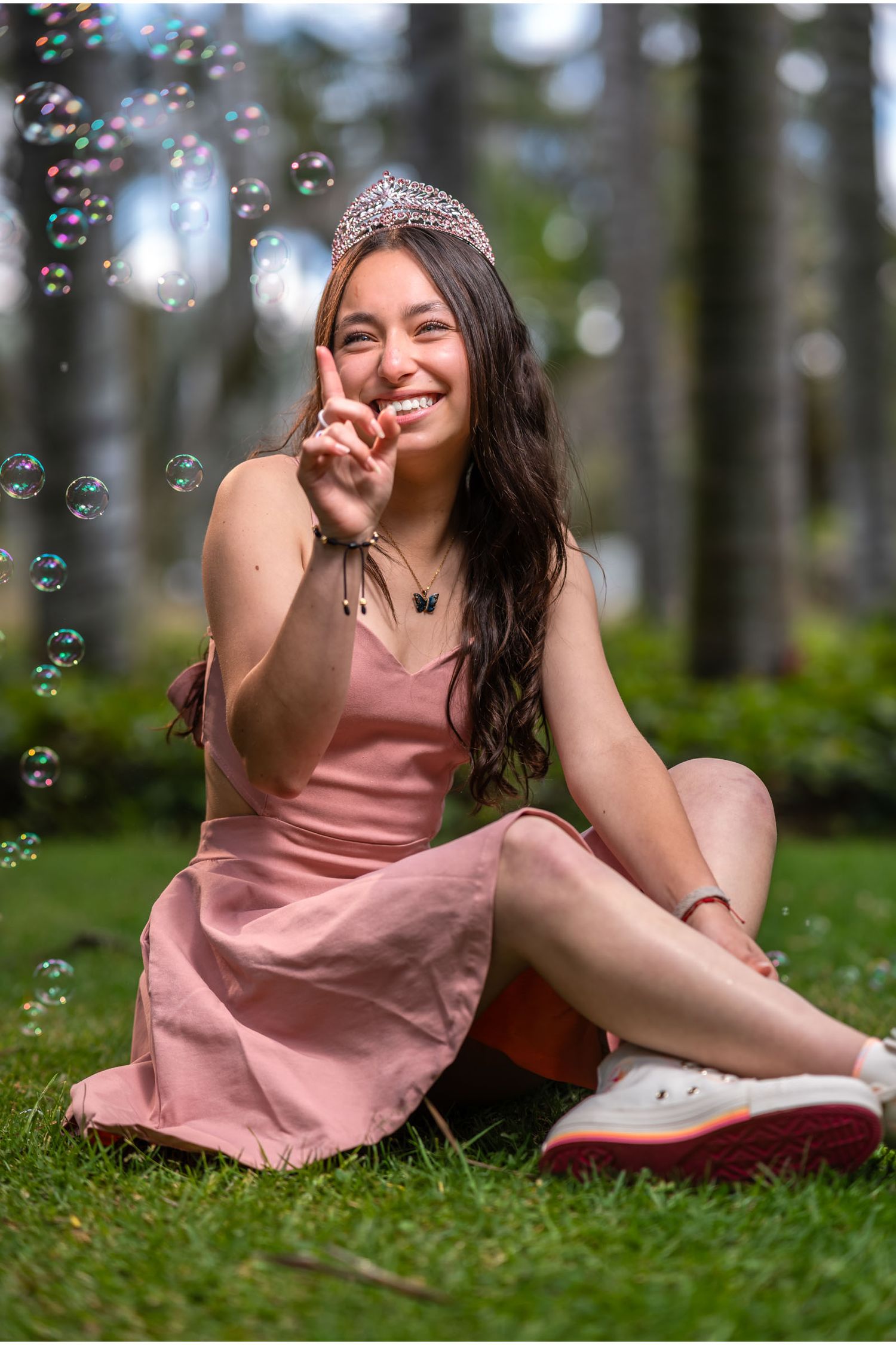 Joven quinceañera sonriente con vestido rosa y tiara sentada en césped, apunta a burbuja flotante en parque.